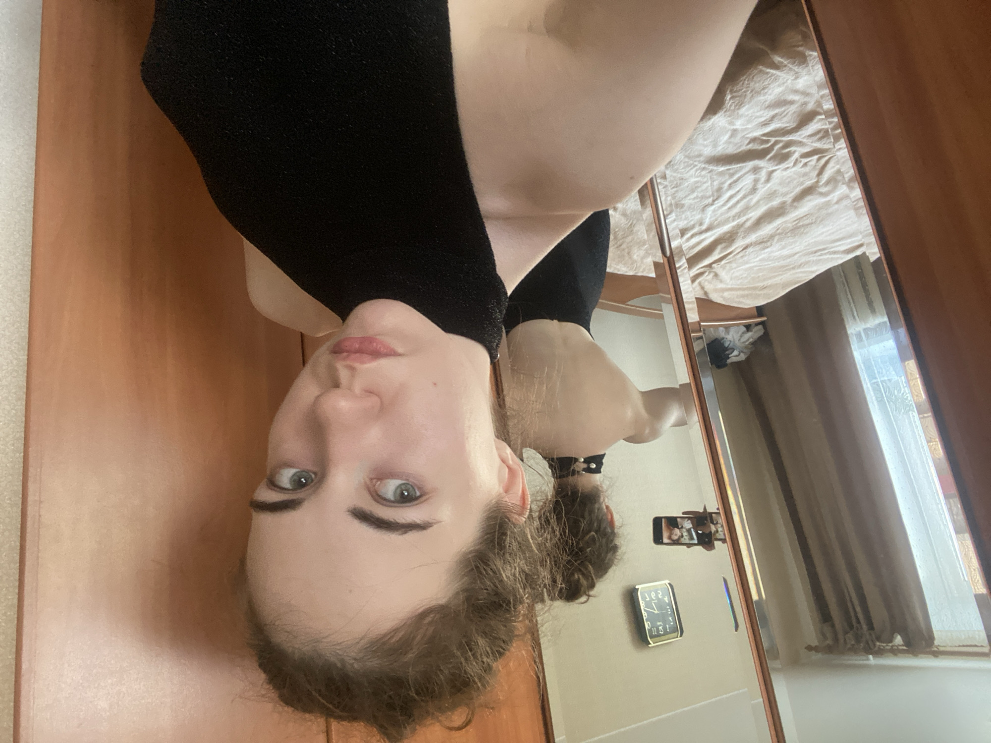 A young woman is captured in a reflection in a mirror, positioned upside down in the frame. She is wearing a black top with a textured finish. Her face is the primary focus, with her light-colored eyes looking directly at the viewer. Her eyebrows are dark and well-defined, and her lips are painted a soft pink. Her light brown hair is pulled back in a messy bun.
The mirror reflects a bedroom setting. A neatly made bed with light beige bedding is visible in the background. To the right of the bed, a window is covered by a light brown curtain. A square clock with Roman numerals is mounted on the wall, showing approximately 2:30. On the wall below the clock, a smartphone is held up, presumably taking the picture, showing another reflection of the woman and the room. The woman appears calm and relaxed, with a neutral expression. The lighting suggests it is daytime, possibly late morning or early afternoon. The scene is indoors, in what appears to be a residential bedroom. There is no visible text other than the numbers on the clock face.