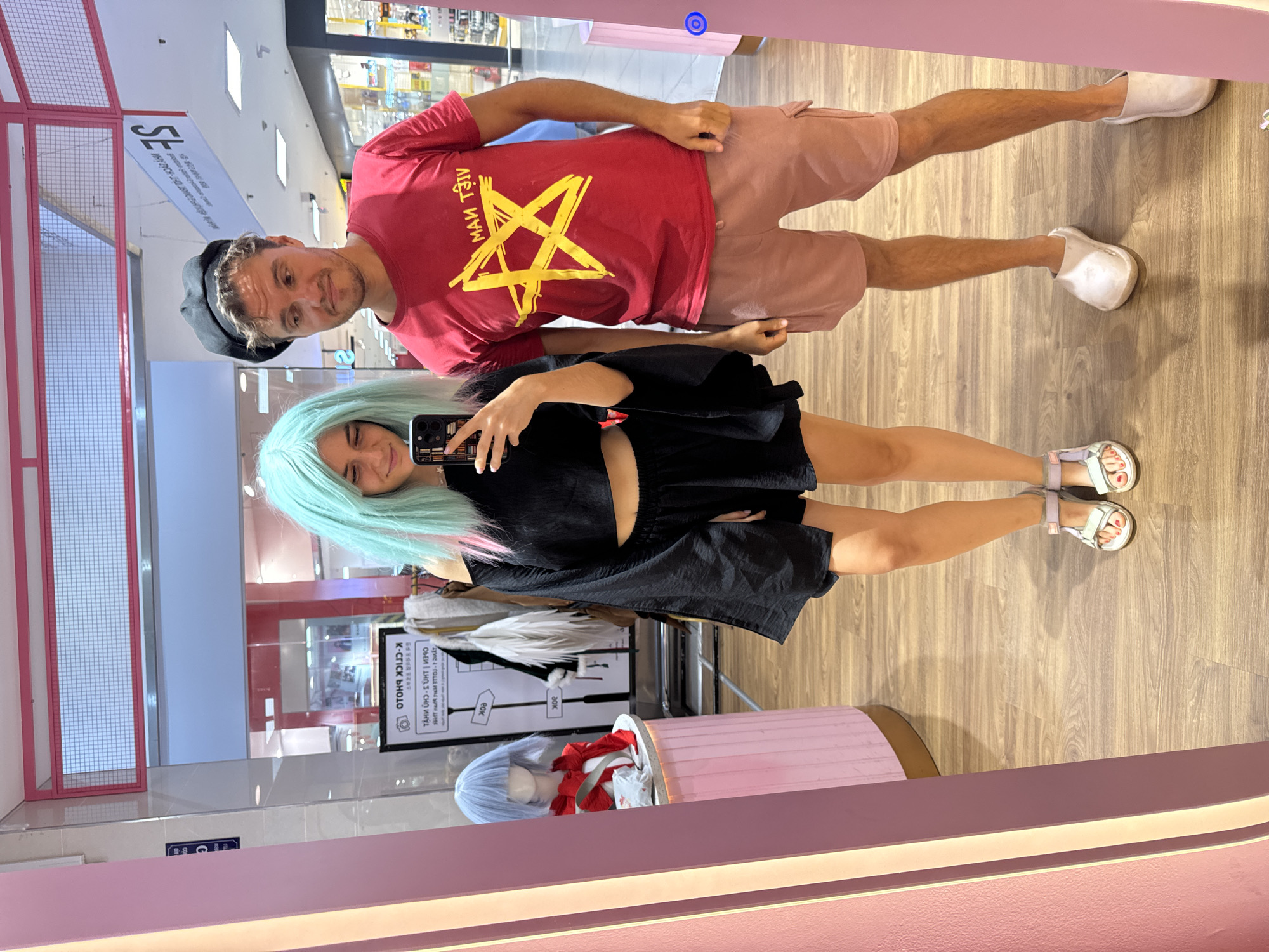A man and a woman are taking a selfie in a store with pink accents and mirrored walls. The man is wearing a red t-shirt with a yellow star design, peach-colored shorts, and white clogs. He also has a dark cap. The woman has light blue hair with pink streaks, a black outfit, and white sandals. The store appears to be selling wigs and cosplay items, as there's a mannequin with a blue wig and a display with a sign reading "K-CLICK PHOTO." The background includes a storefront with a sign that includes "SE" and other text, and wood-look flooring is visible. It’s impossible to confirm if the location is Phú Thủy, Vietnam, based on the image alone.