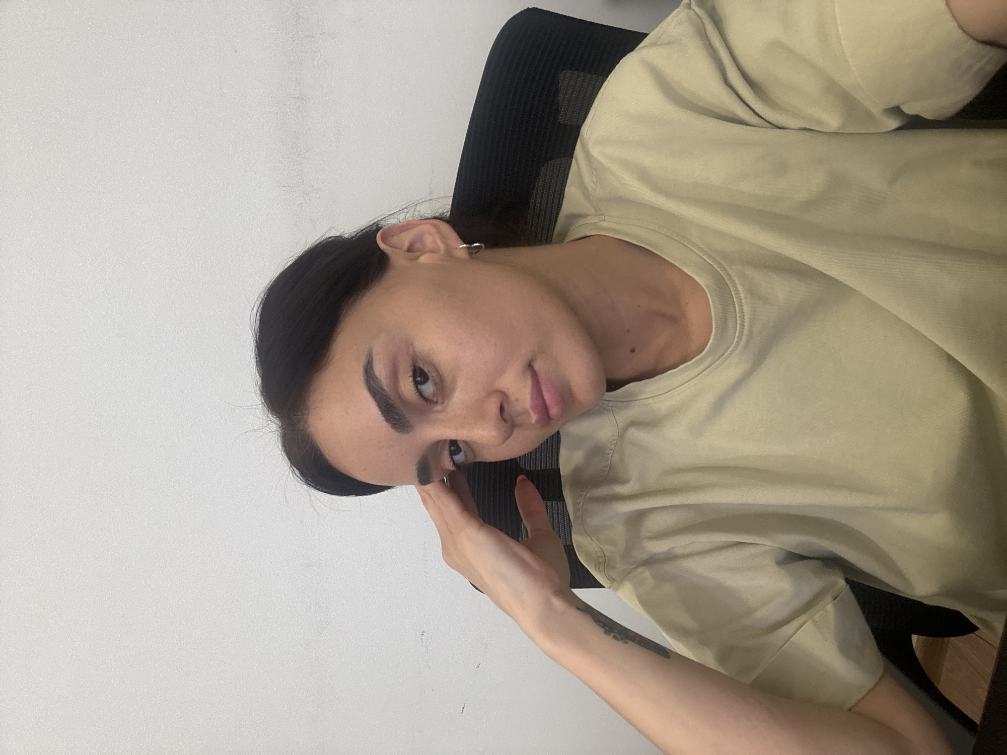A young woman is seated in an office chair, resting her left hand on her forehead. She is wearing a light beige t-shirt and has a small hoop earring in her left ear. A tattoo is visible on her left arm. The background is a plain white wall with some faint marks. The scene appears to be indoors, likely in an office setting, in Tbilisi, Georgia.