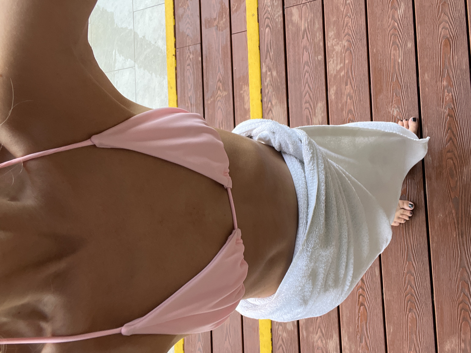 A woman wearing a pink bikini top and a white towel wrapped around her waist stands on a wet wooden deck. The deck has yellow safety stripes. In the background, there is a tiled area that appears to be near a pool. The image was taken in Nha Trang Ward, Vietnam.