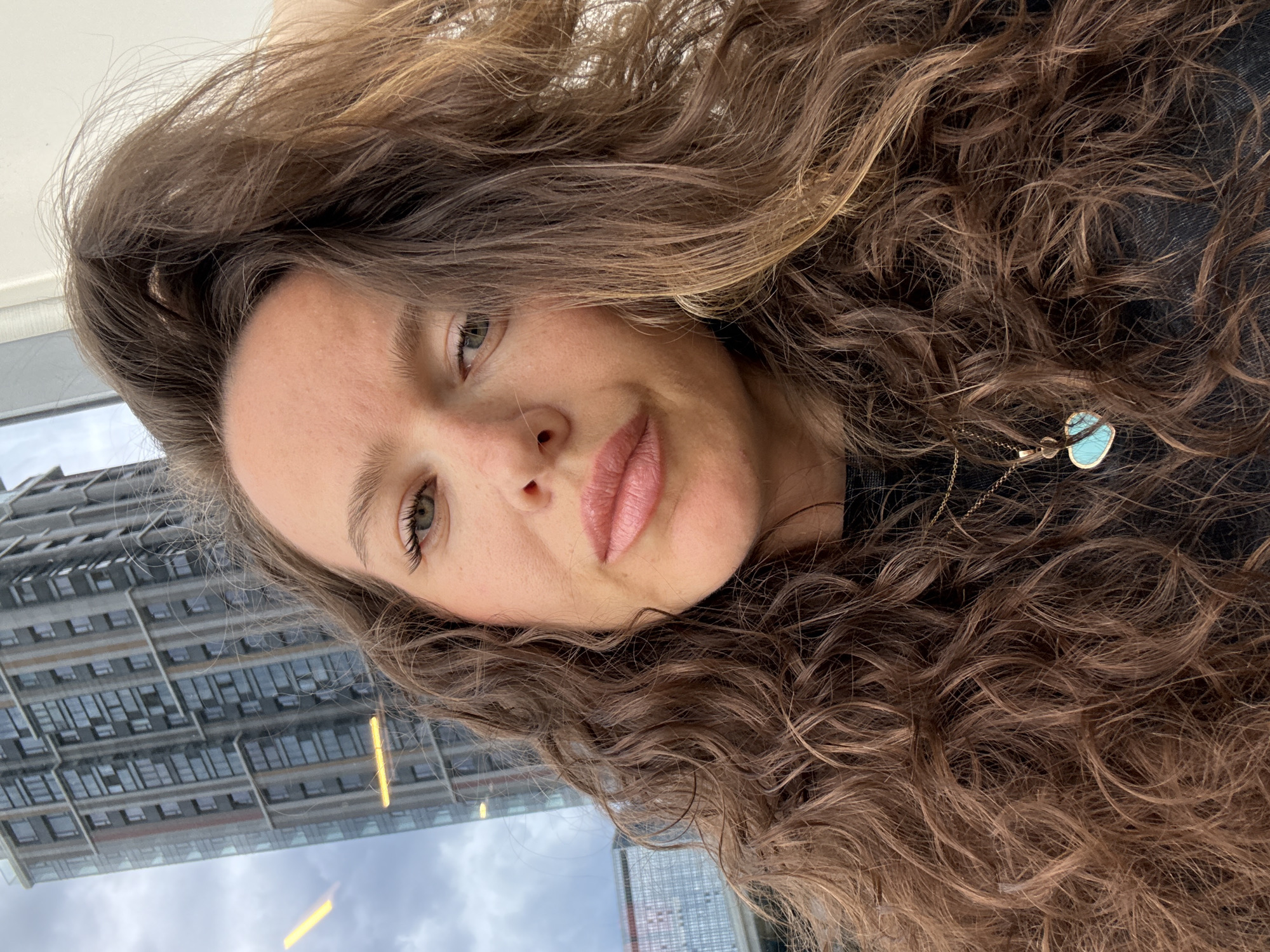 A woman with curly brown hair is taking a selfie. She has light skin, hazel eyes, and pink lips. She is wearing a black top and a necklace with a turquoise pendant. In the background, a reflection of a building with a repeating pattern of windows is visible. The scene is likely inside a building looking out a window. This building could be in the city of Moscow, Russia.