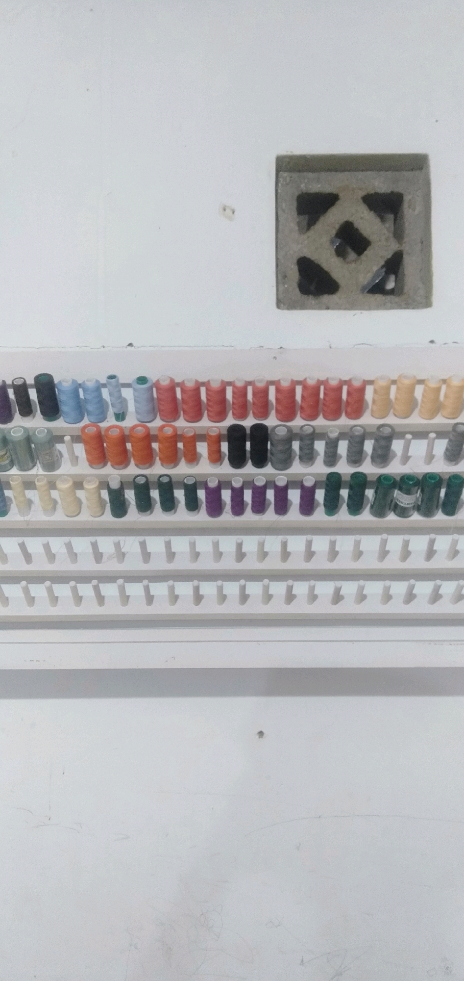 The image displays a white wall with a square vent. Below the vent, a white rack holds rows of thread spools. The spools are organized by color and size, with visible rows of blue, green, orange, pink, yellow, gray, and purple threads. The rack is mounted on the wall and appears to be made of plastic. The lighting suggests an indoor setting. No people or actions are depicted in the image. The location in Cikoneng, Indonesia, is not discernible from the visual information provided.