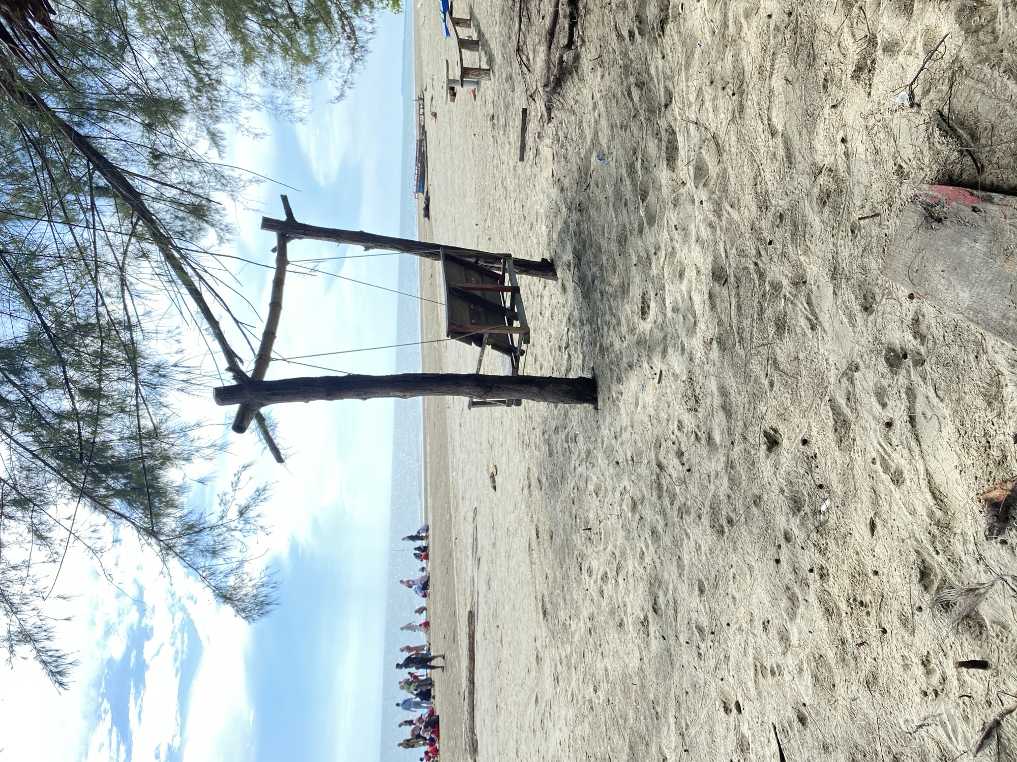 The image depicts a beach scene in Seuneubok Teungoh, Indonesia. In the foreground, there is a makeshift wooden structure with a partially collapsed platform. A pine tree with sparse branches is visible on the left side. The beach itself consists of light-colored sand with visible footprints and debris. In the mid-ground, a group of people is gathered near the water's edge. The ocean and sky are visible in the background. The sky is blue with scattered clouds.