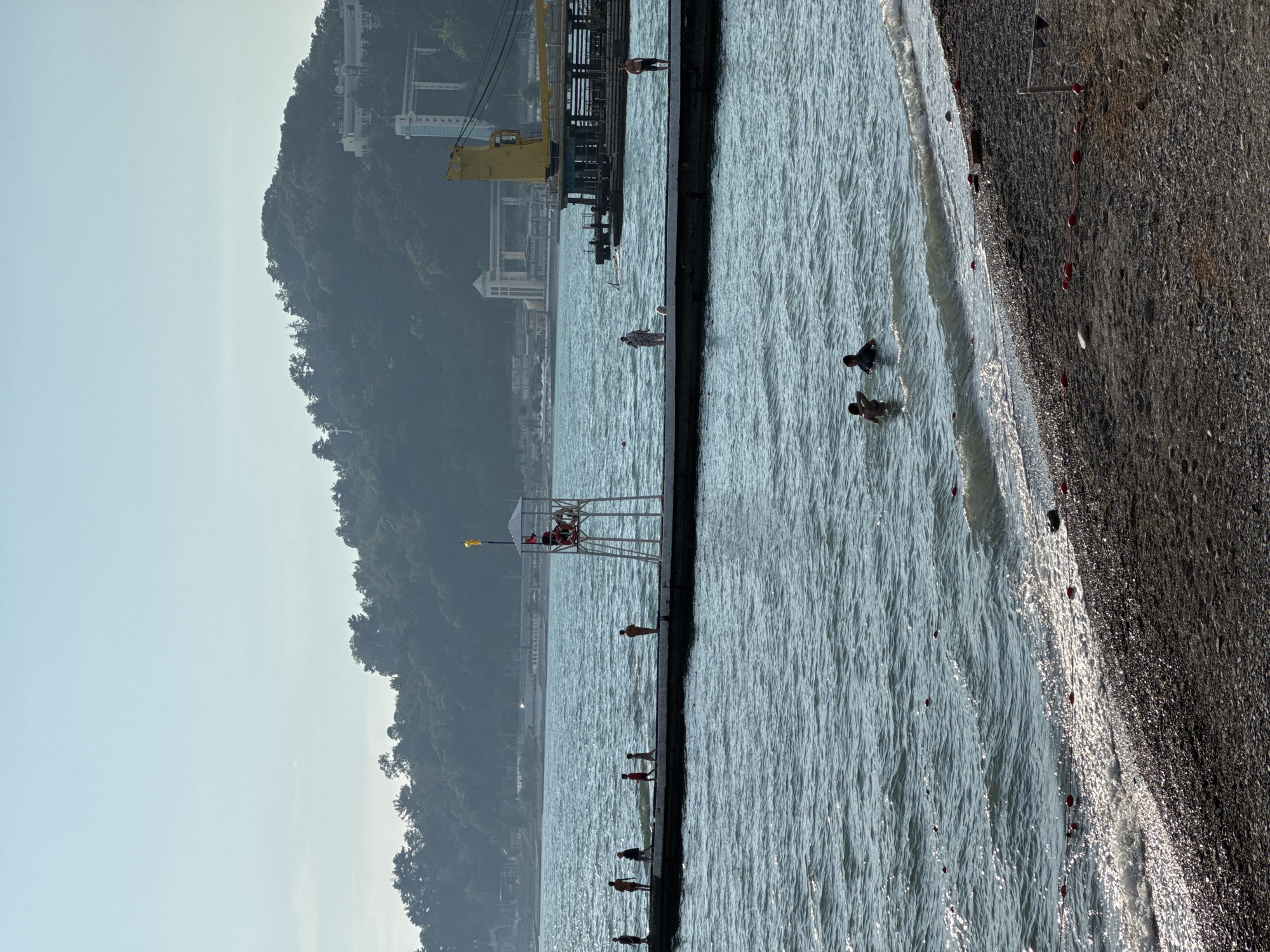 This is a photograph of a beach scene in Sochi, Russia. In the foreground, a pebbly beach is visible with a few red buoys marking the water's edge. The sea is a calm blue with gentle waves lapping at the shore.
Further out, a long concrete pier extends into the water. Several people are seen walking and standing on the pier. On the pier, there is a lifeguard tower with a lifeguard on duty.
In the middle ground, two people are swimming in the sea. In the background, a lush green, tree-covered hill rises steeply. On the hillside, there are some buildings, including a white building with a modern design and a tall, light-colored structure that appears to be a lift or observation tower. A large yellow crane is also visible on a pier or platform in the water, suggesting ongoing construction or port activity. The sky is clear and bright blue.