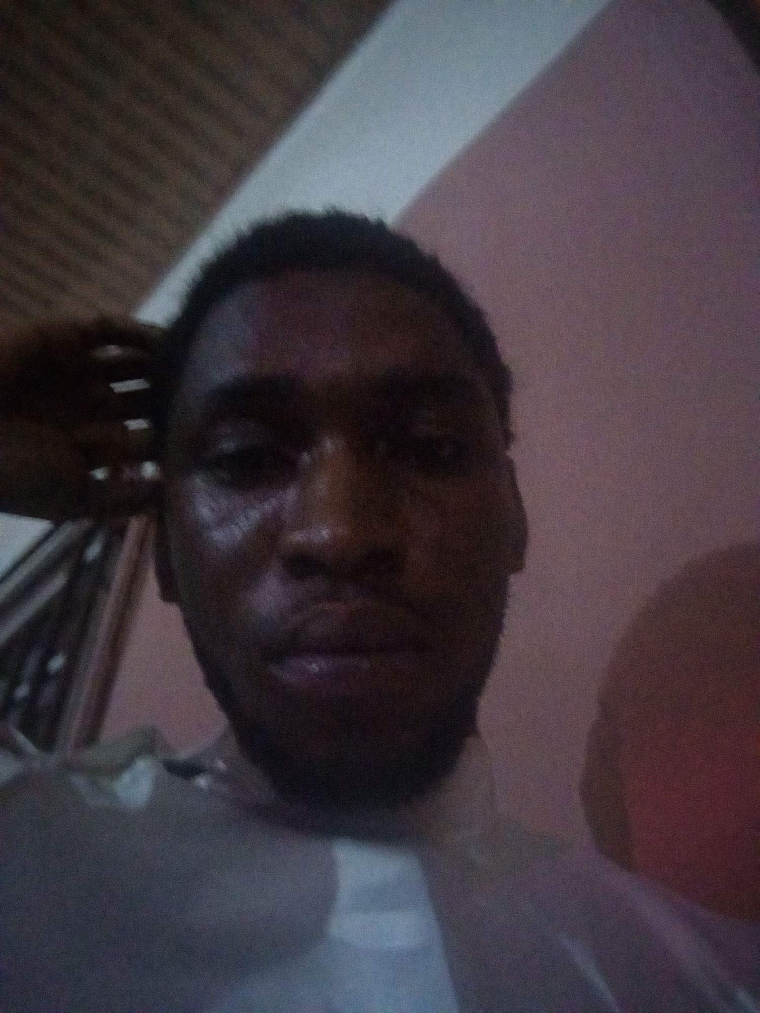 The image shows a close-up self-portrait of a Black man with dark hair and a short beard. His face appears sweaty. He is wearing a light-colored shirt. The background is blurry, showing a wall with a pinkish hue on the right and what appears to be a ceiling with a grid-like pattern on the upper left. A hand is partially visible near his left ear. There is insufficient information to determine a specific location within Lokoja, Nigeria.