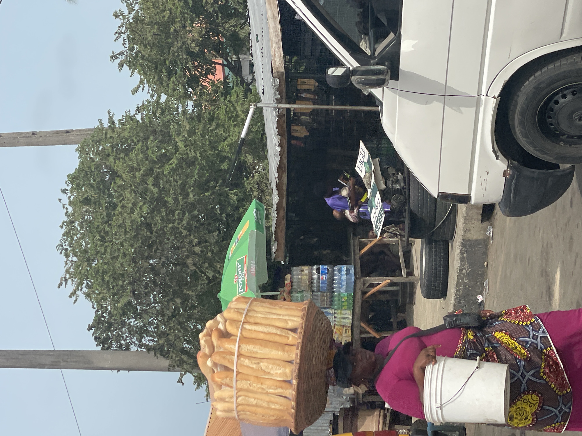In Ajara, Nigeria, a woman is carrying a large basket of bread on her head and a white bucket in her right hand. She wears a magenta shirt and a colorful patterned fabric draped over her shoulder and body. Behind her, there's a roadside setup with a green umbrella labeled "POWER", stacks of bottled water, and other merchandise. To the right, a white van with a black wheel and side mirror is partially visible. In the background, there are trees and a utility pole against a pale sky. A sign reading "EAGLE" is visible near the van.