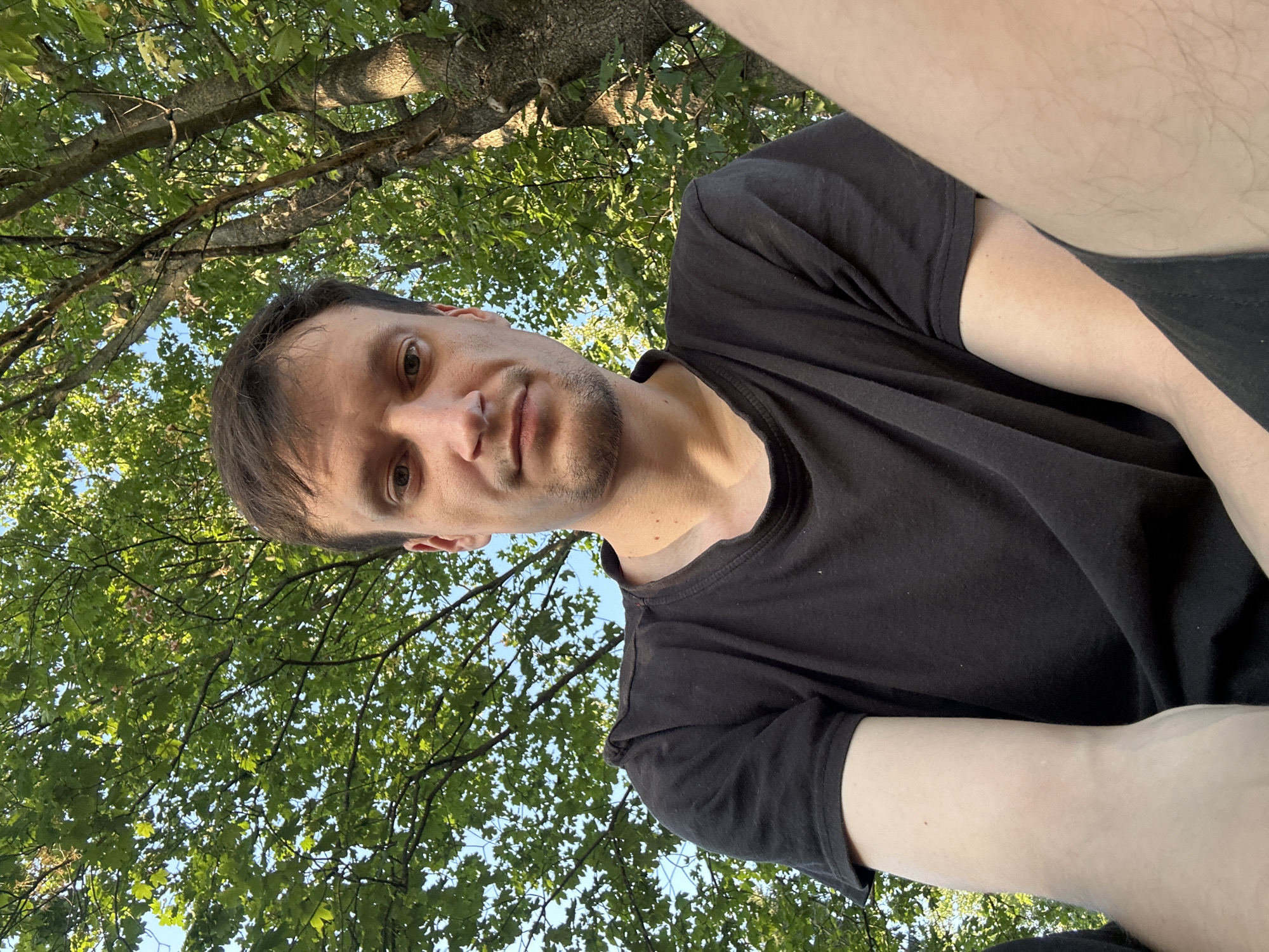 A low-angle, close-up selfie shows a man in his late 20s to early 30s outdoors. He has light brown hair, fair skin, and is wearing a black t-shirt. He is looking directly at the camera with a neutral expression. The background is dominated by the lush green foliage and branches of a large tree, suggesting a park or a wooded area. The sunlight filters through the leaves, creating dappled light and shadows. The location is identified as Makiv, Ukraine.