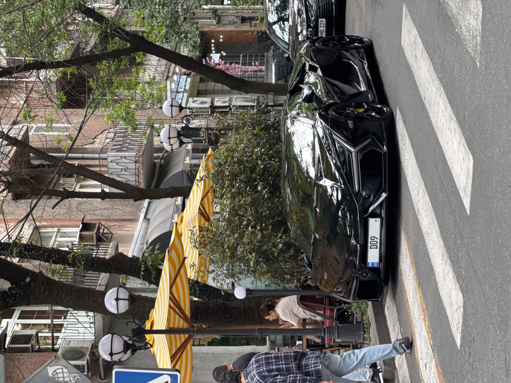 A street scene in Yerevan, Armenia shows a black Lamborghini parked on the side of the road, partially on a zebra crossing. A black car, with a license plate partially visible as "MB 229," is parked behind the Lamborghini. A person in a plaid shirt and jeans walks in the foreground, and another person bends near a sidewalk cafe area with yellow and white striped umbrellas. The license plate of the Lamborghini reads "D09" and includes the Georgian flag. Buildings line the street, and trees with hanging spherical lights are visible. Air conditioning units are mounted on the facades of some buildings.