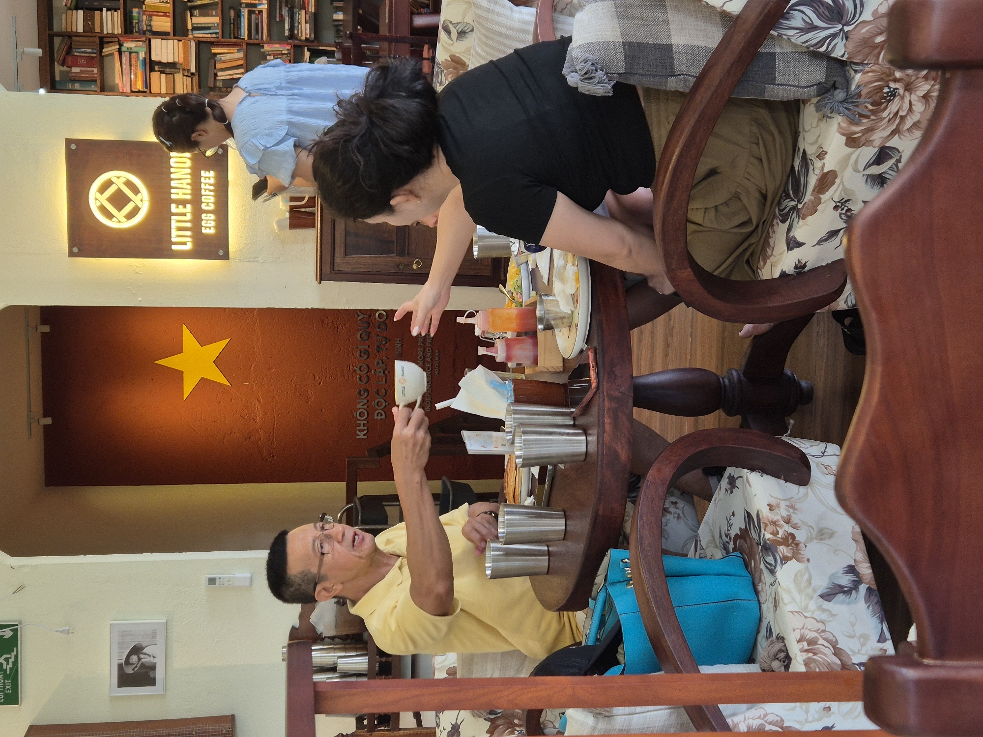 The image depicts an indoor scene within a cafe, identified as "LITTLE HANOI EGG COFFEE" in Da Nang, Vietnam, suggested by a sign on the wall. Two individuals are seated at a round wooden table. A man, wearing a yellow polo shirt and glasses, is offering a cup to a woman who is also seated at the table. The woman, wearing a black t-shirt, reaches out to accept the cup. The table is laden with various items, including several metal cups, plates with food, condiments in small bottles, and a napkin. A blue tote bag rests on the floor near the man. The cafe's interior is decorated with a large red wall featuring a prominent yellow star, bookshelves filled with books, and framed pictures on the wall. The chairs are upholstered with a floral pattern. The overall atmosphere appears to be casual and convivial.