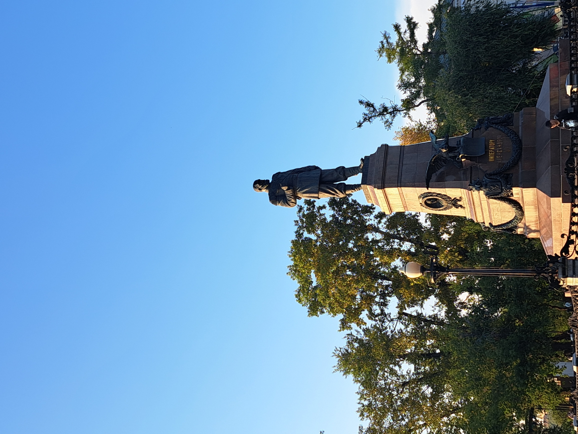The image depicts the Monument to Alexander III in Irkutsk, Russia, situated in front of a clear blue sky. The monument is a tall, stone structure with a statue of a man in military attire at the top. The base of the monument features intricate carvings and inscriptions. In the foreground, there is a lamppost and a black metal fence. Lush green trees are visible behind the monument, suggesting a park or public square setting. A few people are present, including one individual leaning on the fence near the bottom right corner of the image. The lighting suggests it is daytime, with sunlight illuminating the scene.