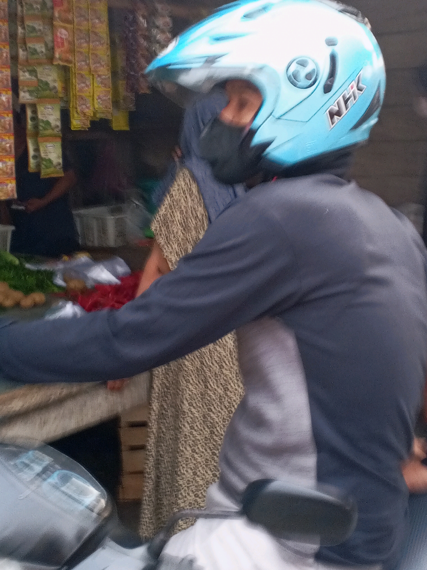 In Kadikaran, Indonesia, a person wearing a light blue motorcycle helmet with "NHK" branding rides a motorcycle. The helmet obscures most of their face, revealing only the bridge of their nose and an eye. The person is wearing a dark long-sleeved shirt. They are seen from the shoulder down, with the motorcycle's handlebars and dashboard visible in the foreground. Behind the rider, a market stall displays various packaged goods hanging on the wall and fresh produce, including red items and what appear to be potatoes, on a table. The environment suggests a busy street market.