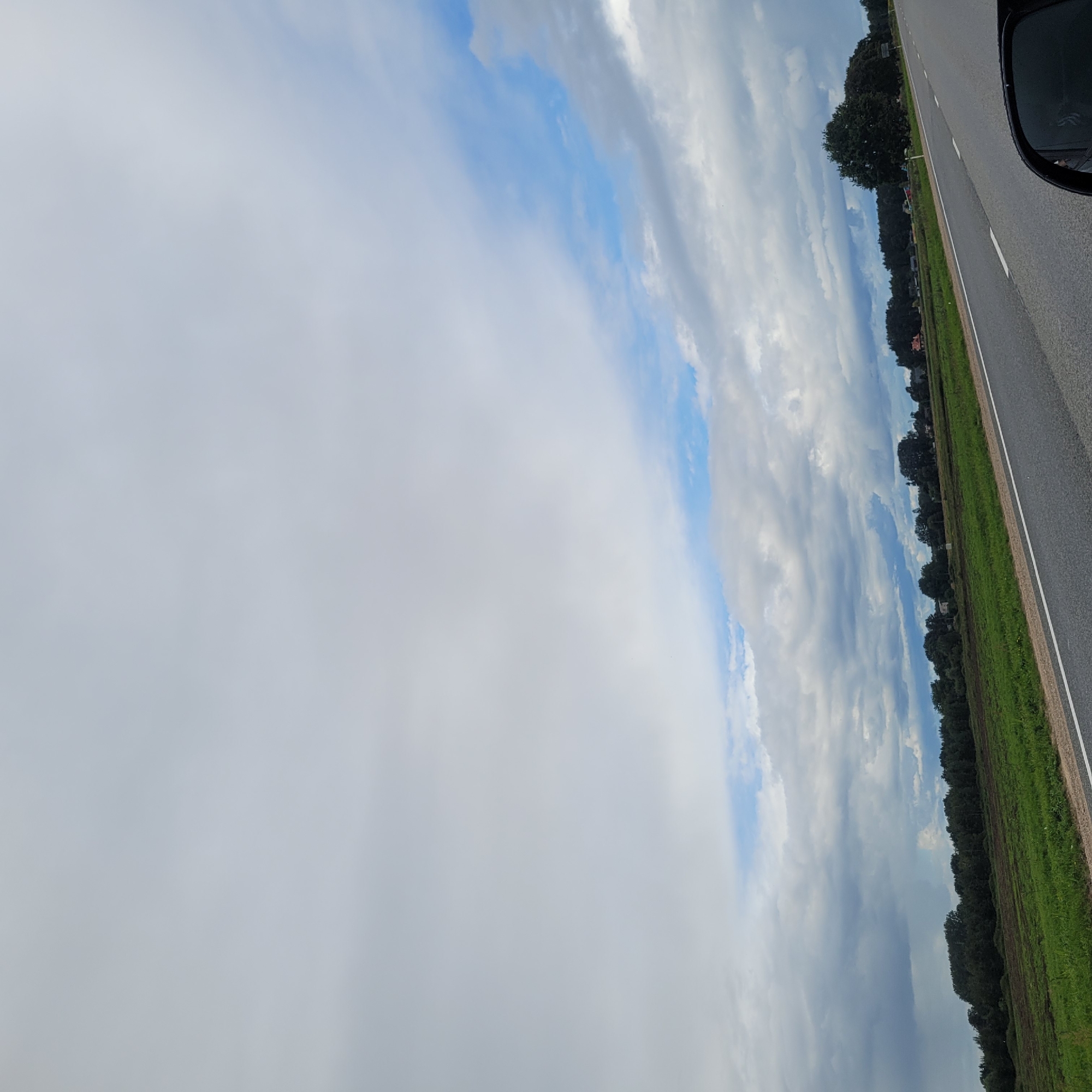 The image depicts a view from a vehicle traveling on a paved road, likely in or near Jaunmārupe, Latvia. The road runs from the lower right to the upper left of the frame. To the left of the road, there is a green field, which appears to be farmland, with some areas showing tilled soil. Beyond the field, a dense line of trees forms the horizon, with some buildings, possibly houses or farm structures, visible interspersed within or behind the treeline. The sky is predominantly filled with clouds, with patches of blue sky visible towards the upper right. The clouds are varied in density and color, ranging from white and fluffy to darker, more ominous formations. The overall lighting suggests it might be overcast or nearing dusk. A side mirror of the vehicle is visible in the lower right corner of the image, reflecting a portion of the sky and the surrounding environment.