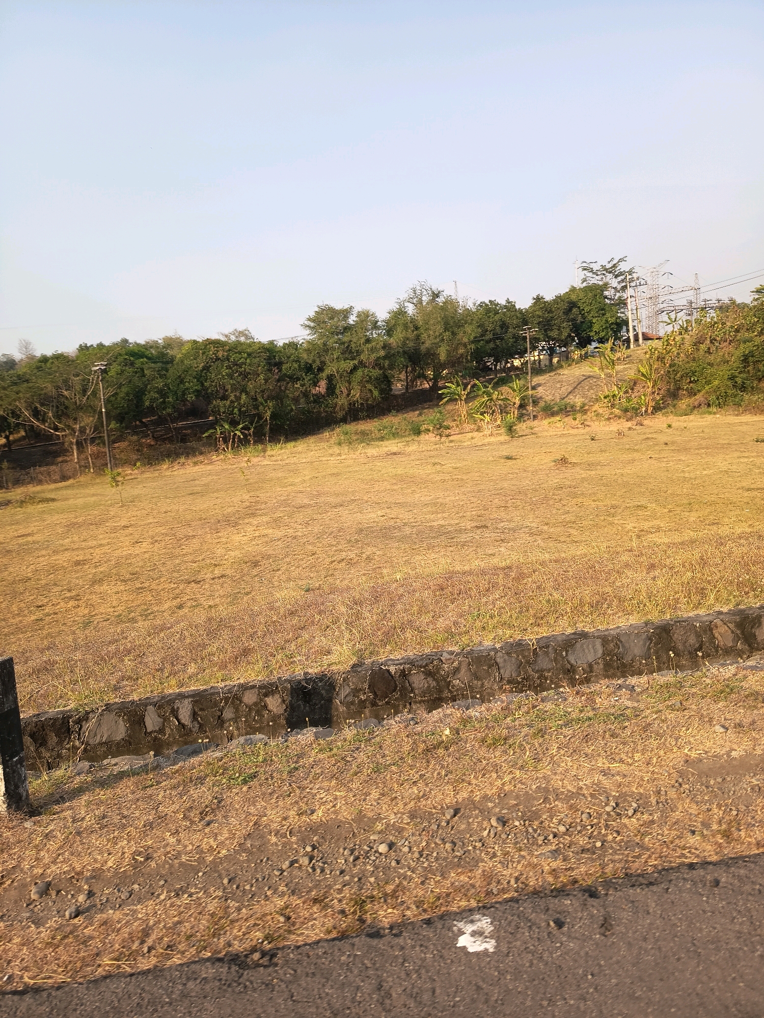 The image depicts a dry, grassy field in Kalangbancar, Indonesia, with a low stone wall in the foreground. Beyond the field, a line of trees separates the open area from a section of industrial infrastructure, characterized by tall power lines and transmission towers. The sky above is clear and a pale blue, suggesting a sunny day. The overall impression is one of a rural or semi-industrial landscape under a bright sky.
