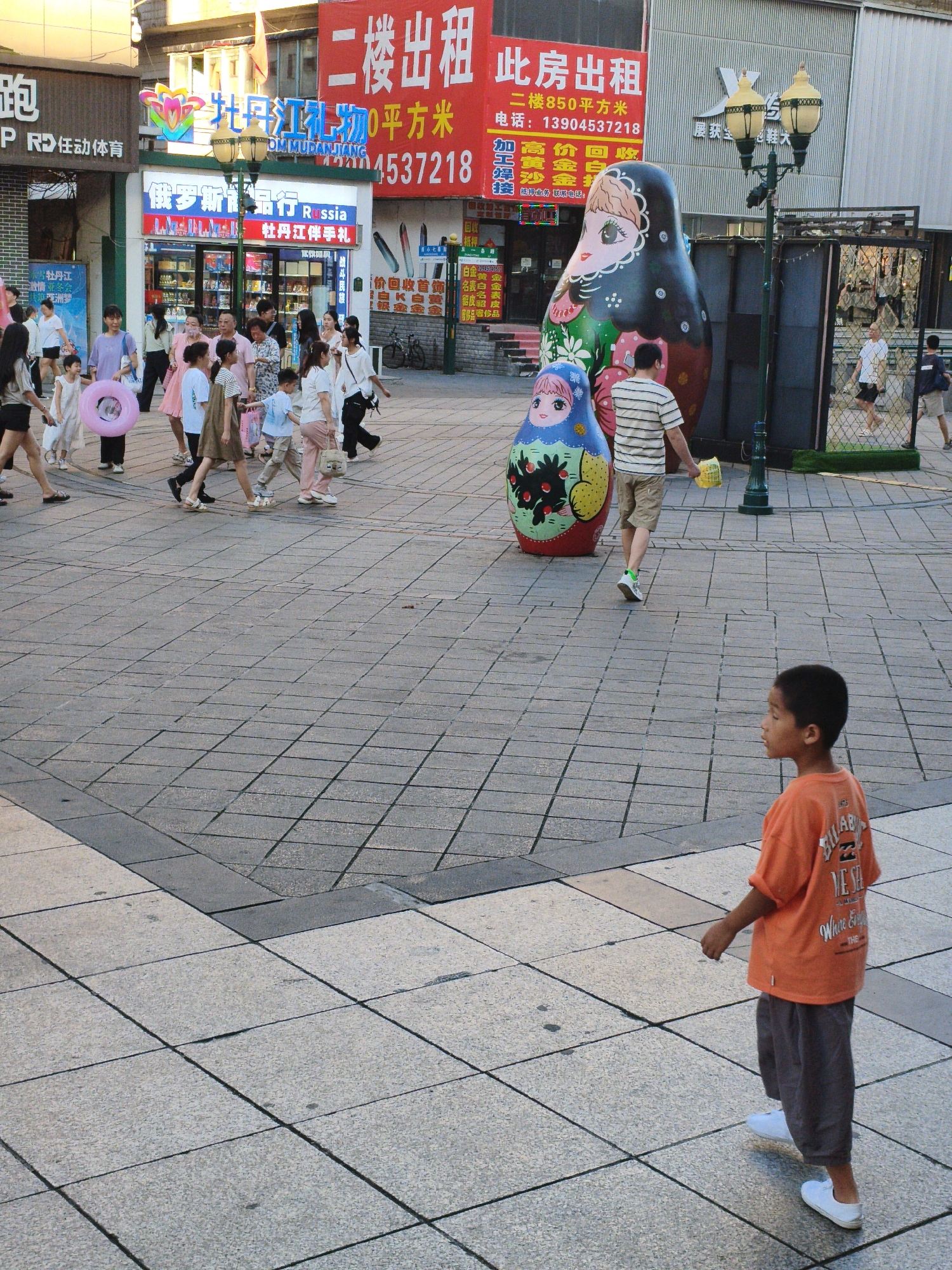In Mudanjiang, China, a street scene unfolds on a paved plaza under diffused sunlight. Multiple people are present, including adults and children, some walking in groups and others individually. A young boy in an orange t-shirt and grey pants walks away from the camera. In the background, large, decorative nesting dolls are displayed. Various shops with signs in Chinese are visible, one advertising "Mudanjiang Gifts." A lamppost with ornate lighting stands near the nesting dolls. The ground is tiled with a pattern of squares.