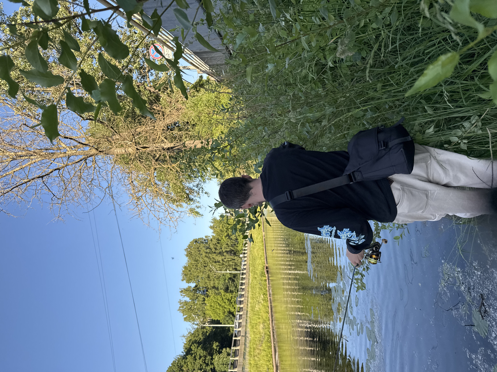 The image depicts a young man fishing in a narrow canal bordered by dense green vegetation and a concrete embankment. He is facing away from the camera, holding a fishing rod and reel. He wears a black long-sleeved shirt with light blue graphics on the sleeves and light-colored trousers. A dark messenger bag is slung over his right shoulder. The canal water is dark and reflects the surrounding trees and a bright blue sky with a few wispy clouds. On the far bank of the canal, a grassy slope leads up to a road with streetlights and traffic visible in the distance. The setting appears to be a canal in Saint Petersburg, Russia, likely within a park or along a waterway connected to the city's extensive network of canals and rivers. The time of day is likely late afternoon or early evening, given the quality of light.