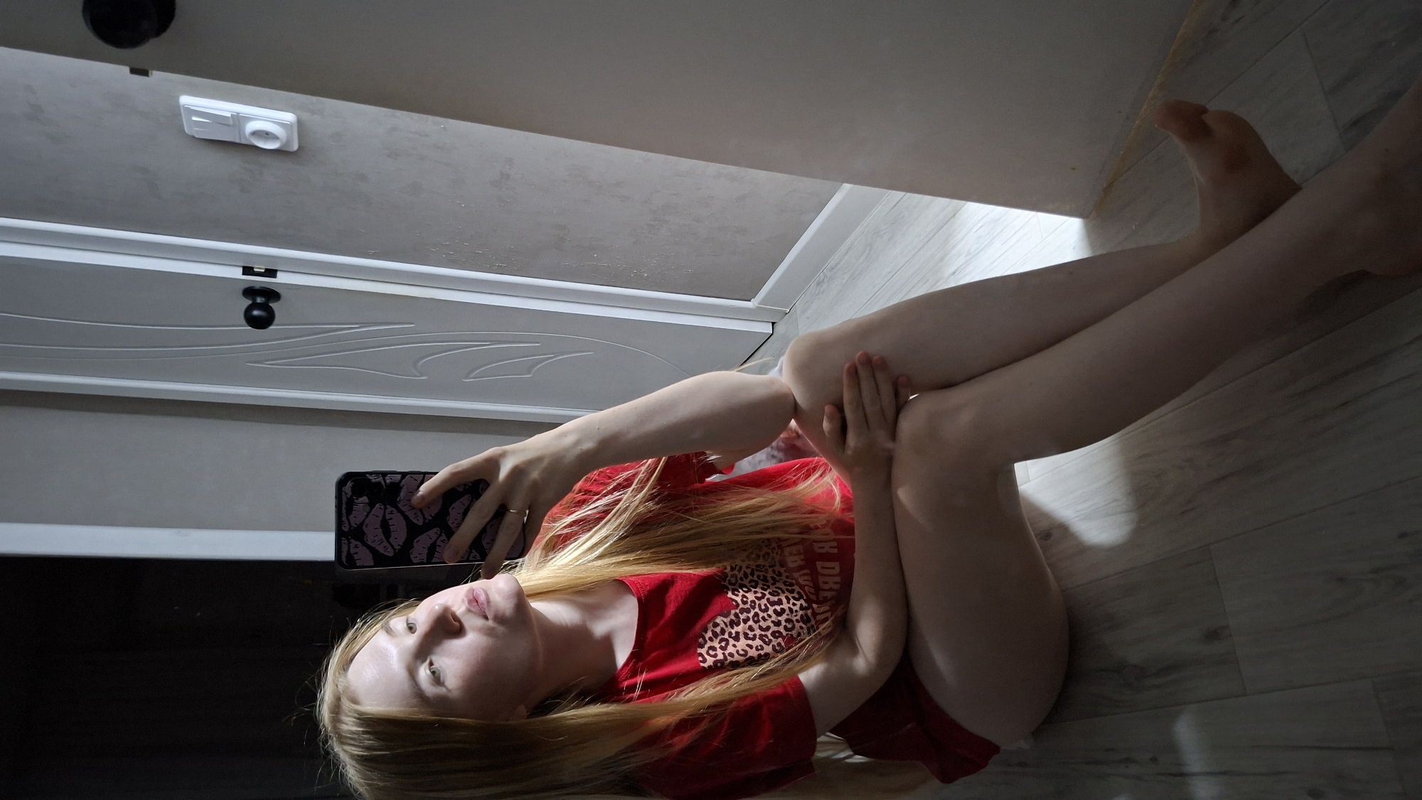 A young Caucasian woman with long blonde hair and fair skin is sitting on a wooden floor, taking a selfie with her phone. She is wearing a red t-shirt with a leopard print heart graphic on it. The woman is looking at her phone, with her left hand holding it and her right hand resting on her knee. Her legs are bent at the knees, with her right leg crossed over her left. The background of the image shows a white door with a black doorknob, a light-colored wall, and a part of what appears to be a mirror. The lighting suggests the photo was taken indoors, possibly in a home. The scene is devoid of any urban or specific city landmarks.