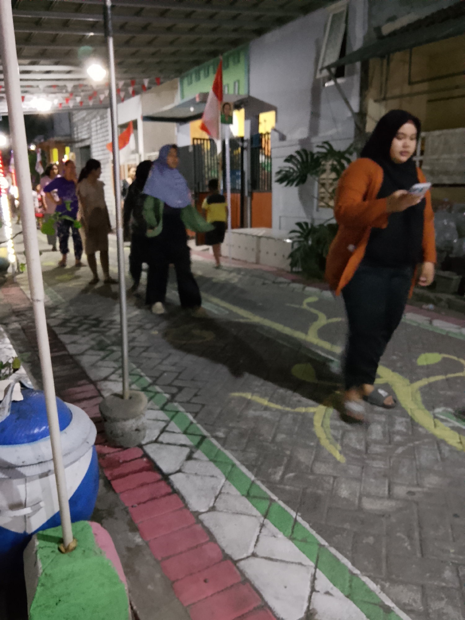 The image depicts an outdoor scene at night, likely on a street or public walkway in Banjarsari, Indonesia. Several individuals are visible, including a woman in the foreground wearing a black hijab and an orange cardigan, holding a smartphone. Other people are seen walking and standing further down the path, some dressed in casual attire. The environment features a paved walkway with decorative patterns in yellow, pink, and white. Red and white triangular flags are strung overhead, suggesting a celebratory or nationalistic event. Buildings with windows and doors line the background, and a large blue and white container is present on the left side of the frame. A pole with a concrete base stands near the container, and another similar pole is visible in the mid-ground. The lighting is dim, consistent with an evening setting, with artificial lights illuminating parts of the scene.