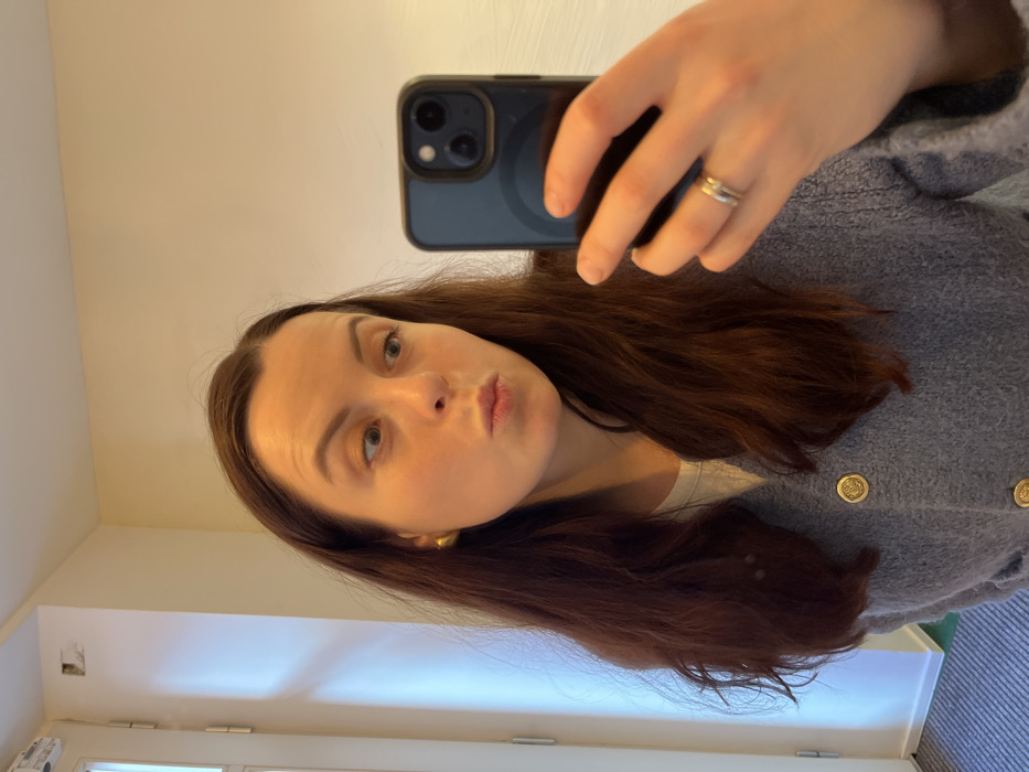 A female subject is depicted taking a mirror selfie indoors. She has long, dark brown hair and fair skin. Her attire includes a white undershirt beneath a textured, light grey cardigan fastened with large, circular gold buttons. She wears gold spherical stud earrings and a ring, appearing as two stacked bands, on the ring finger of her left hand. The subject holds a dark-colored smartphone, featuring a visible triple camera array, in her left hand, positioned to capture her reflection. Her lips are pursed in a pout or kiss, and her eyes are wide, directed towards the phone screen. The background consists of plain, light-colored walls, possibly off-white or light beige. A white door frame with visible metal hinges is present on the far left. A small, dark, rectangular recess or fixture is observed high on the left wall, above the door frame. Natural light illuminates the scene from the left. This scene is located in the city of Kaiserslautern, Germany.