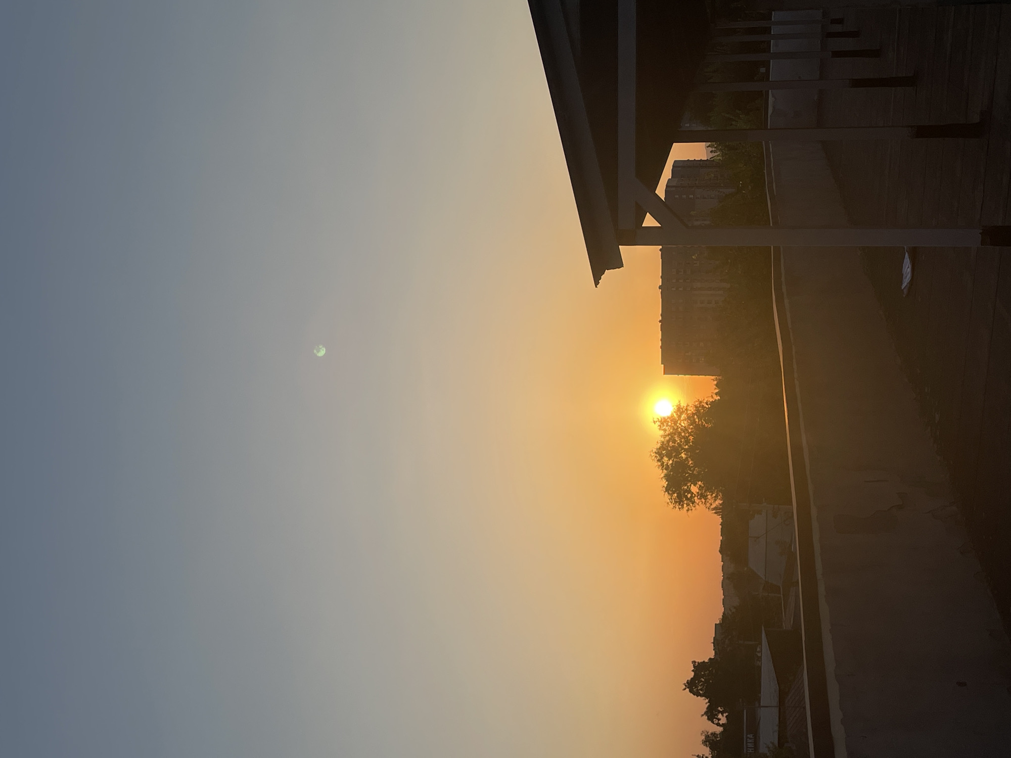 The image captures a serene sunset over the city of Almaty, Kazakhstan. The sky transitions from a soft blue at the top to a warm orange hue near the horizon, indicating the sun is setting. A faint, pale orb, possibly the moon or a cloud illuminated by the setting sun, is visible in the upper portion of the sky.
In the mid-ground, the sun itself is a bright, golden circle, partially obscured by the silhouette of lush green trees. Beyond the trees, several multi-story buildings are visible, their forms softened by the warm evening light. To the right of the image, a covered walkway or a rooftop structure with white pillars and a dark roof dominates the foreground. The surface below the walkway appears to be a flat, dark rooftop.
In the lower foreground, a concrete wall, likely part of a rooftop, stretches across the image. The wall shows signs of wear, with peeling paint and visible textures. A white object, possibly a piece of paper or plastic, lies on the dark rooftop in front of the wall.
The overall impression is one of urban tranquility at the end of the day, with the natural beauty of the sunset complementing the city's architecture. The scene is devoid of any visible people or animals.