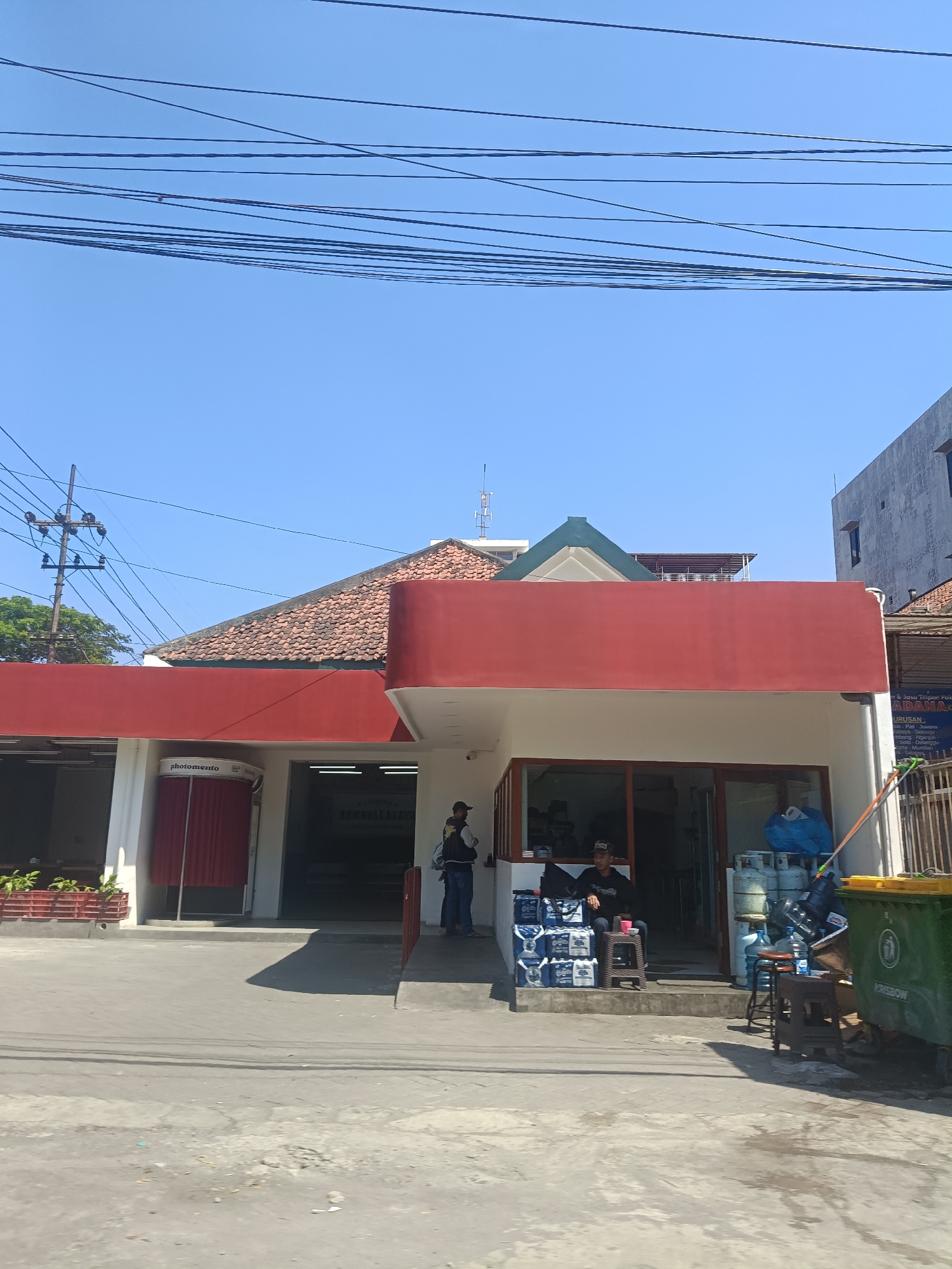 The image shows a street-level view of a business establishment in Surabaya, Indonesia, on a clear, sunny day. The building features a prominent red awning over its entrance. To the left of the entrance, there is a red photo booth with the text "photomento" visible. Inside the building, there is a display of water bottles and gas cylinders. Two men are present, one standing near the entrance and another seated on a stool in front of the display. A green dumpster is located to the right of the building. Overhead, a tangle of electrical wires crisscrosses the sky, typical of urban streetscapes. The street in front of the building appears to be paved with concrete, with some debris visible. The architecture suggests a commercial or service-oriented business, potentially a distributor of water or other goods.