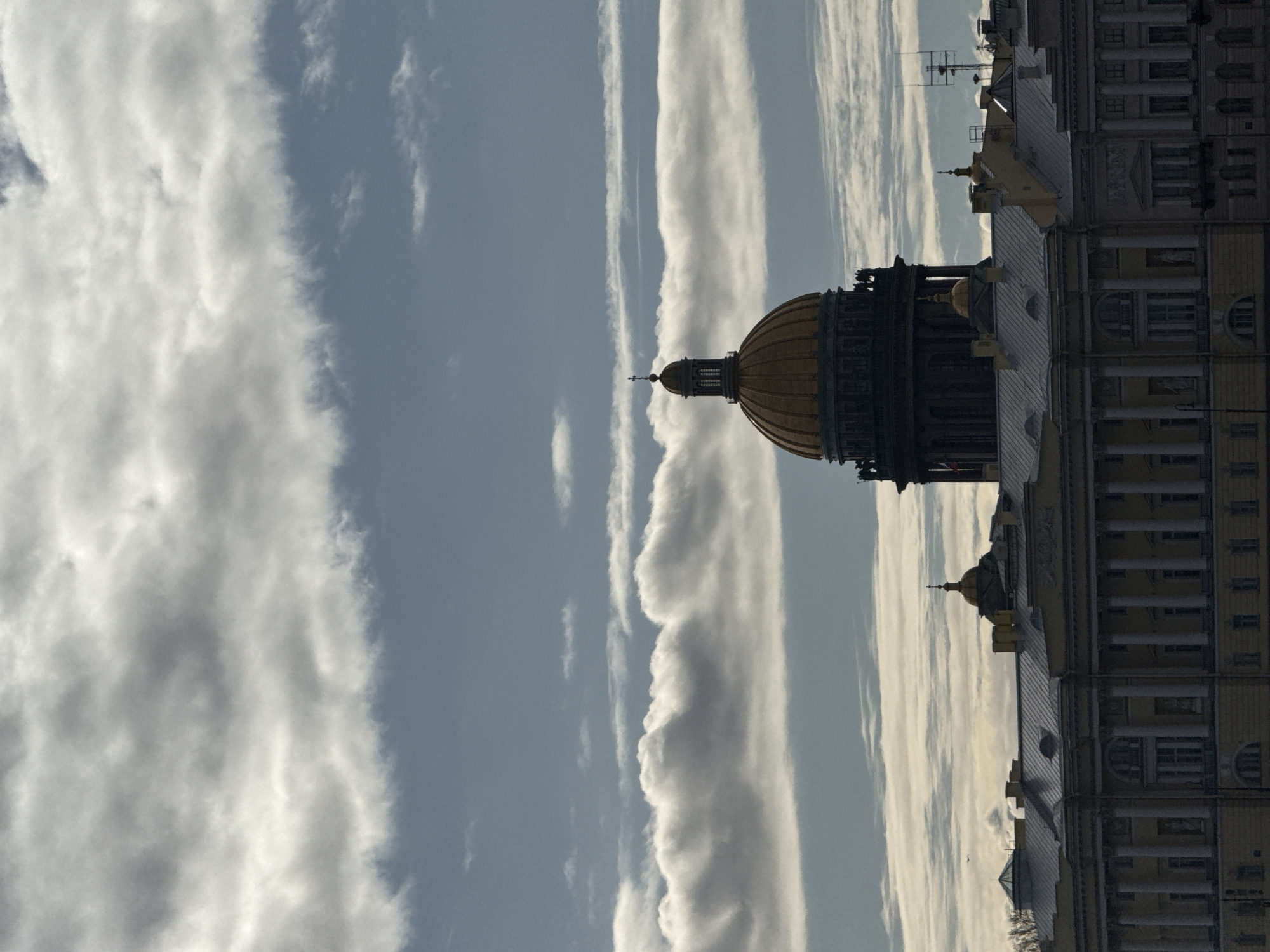 The image, viewed upright, depicts an architectural landscape dominated by a grand golden dome and an adjacent historic building against a partially cloudy sky. The most prominent feature is a large golden dome, likely belonging to Saint Isaac's Cathedral in Saint Petersburg, Russia, situated on the left side of the frame. The dome is ornate, featuring visible ribbed sections, and is crowned with a smaller cupola topped by a cross.
To the right of the dome, the upper levels and rooftop of another substantial building are visible. This structure exhibits classical architectural elements, including multiple columns, evenly spaced windows, and a decorated facade. Its rooftop features several spires, smaller domes, and what appear to be antennae or communication structures.
The sky above the buildings occupies a large portion of the image. It is a light blue-grey, traversed by several long, linear streaks of white clouds, possibly contrails or high-altitude cirrus formations, stretching horizontally across the scene. Interspersed with these lines are broader, more diffused patches of white and grey clouds.
The lighting suggests a soft illumination, characteristic of late afternoon or early morning, with the golden dome reflecting light and parts of the buildings displaying subtle shadows. No people are visible, leaving the focus entirely on the monumental architecture and the atmospheric sky. There is no visible text in the image. The scene conveys a sense of historical grandeur and urban majesty in Saint Petersburg, Russia.