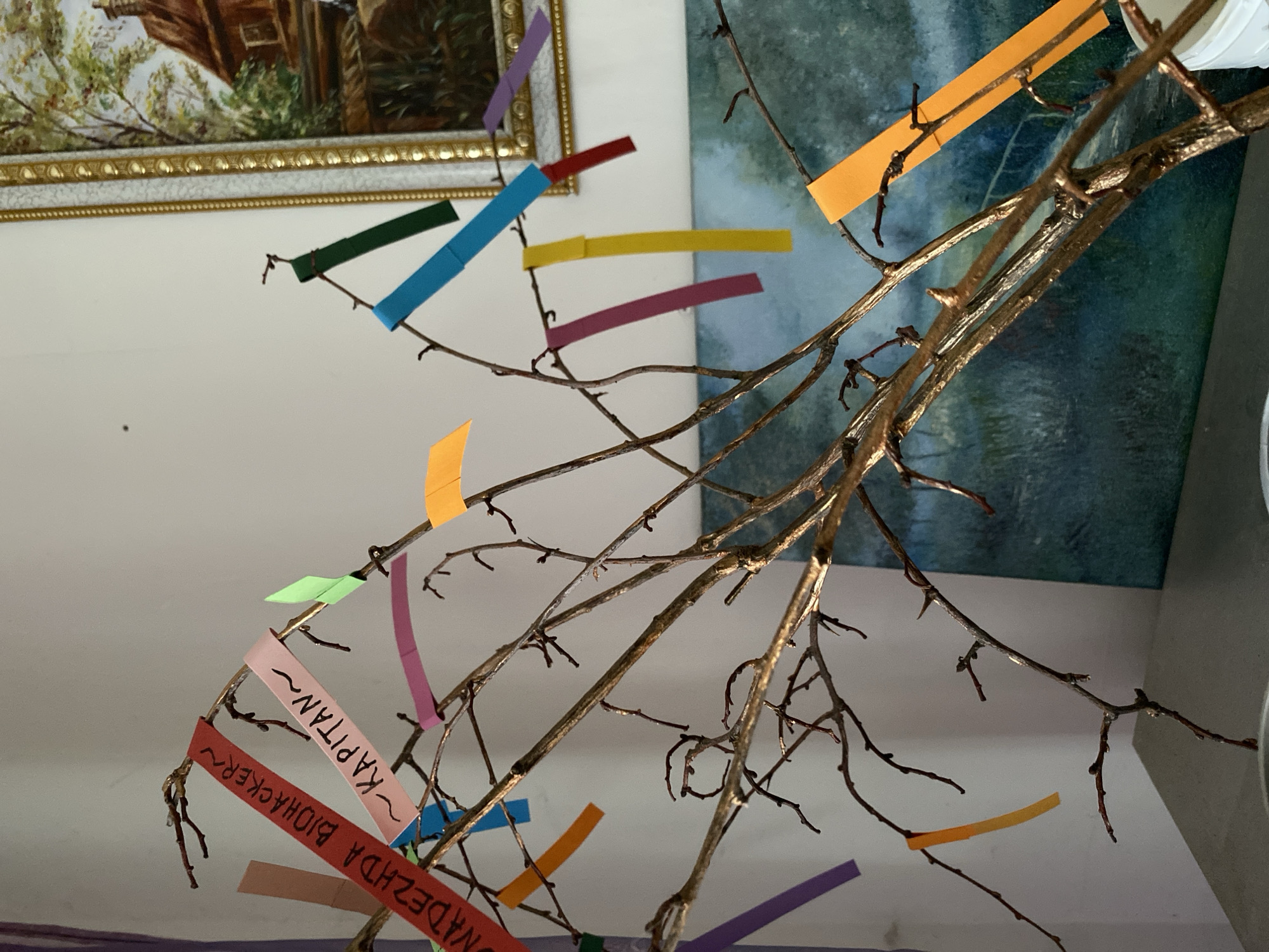 The image shows a close-up of a golden-colored tree branch, likely an artificial decoration, adorned with colorful paper strips. These strips are of various colors including red, pink, green, blue, yellow, orange, and purple. Some of the paper strips have writing on them in Cyrillic script. The red strip on the left reads "NADEZHDA BIOHACKER~" and the pink strip next to it reads "~KAPITAN~". The background consists of a white wall, a framed painting depicting a house and trees, and a large painting or tapestry of a landscape with a body of water. The scene appears to be indoors, likely in a home or office setting in the city of Bílina, Czechia. The lighting is soft, suggesting it might be daytime.