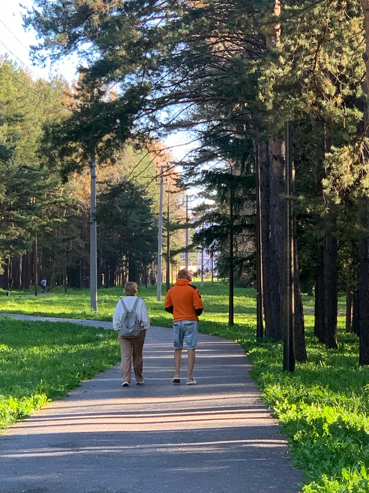 The image depicts a paved pathway through a wooded area, likely a park in Prokopyevsk, Russia. Two individuals are walking away from the camera on the pathway. The person in front, on the left, is wearing a white top with a light gray backpack and light brown pants. The person behind, on the right, is wearing an orange hooded sweatshirt and light blue shorts. The pathway is lined with tall trees, predominantly pine trees, casting shadows across the path. Lush green grass and some smaller plants border the pathway. In the background, more trees and utility poles are visible, suggesting a park or a suburban area. The sky is partly cloudy and a soft, warm light indicates it's likely late afternoon or early morning. A third person is visible in the distance on the left, walking across the grassy area.