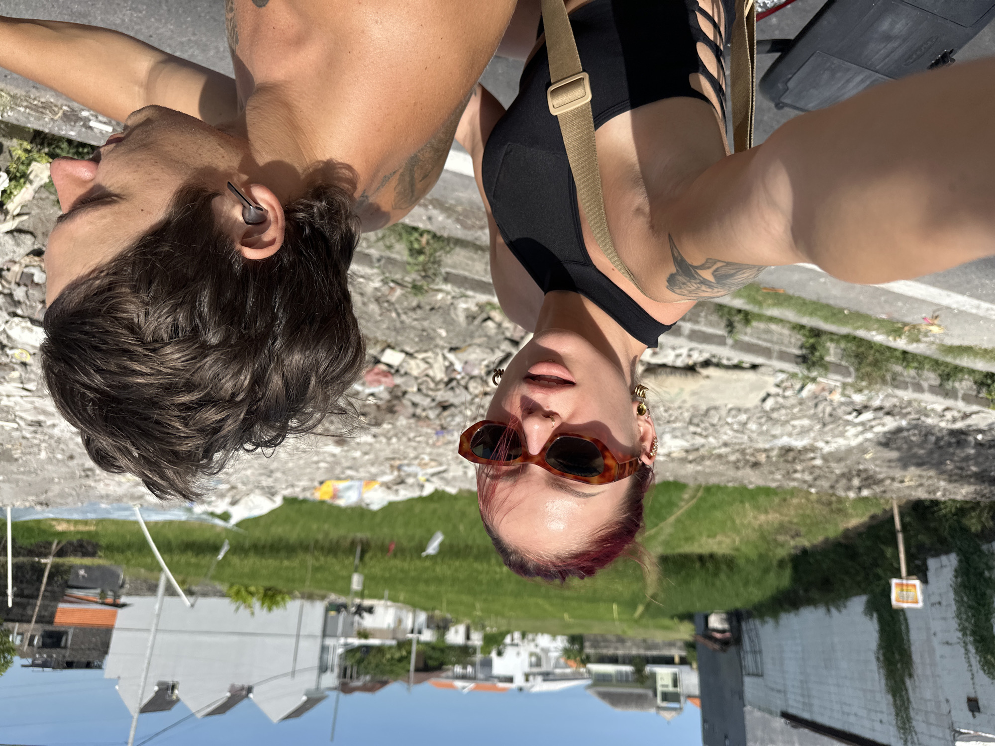 Two people are visible in the photograph, a man and a woman. The man has dark hair and is wearing an earpiece. The woman has dark hair with red streaks, sunglasses, and multiple earrings. She is wearing a black top with cutouts. Both individuals have tattoos on their arms. The background shows a street, grass, and buildings, suggesting an urban environment. Based on the metadata, the location is in North Kuta, Indonesia.