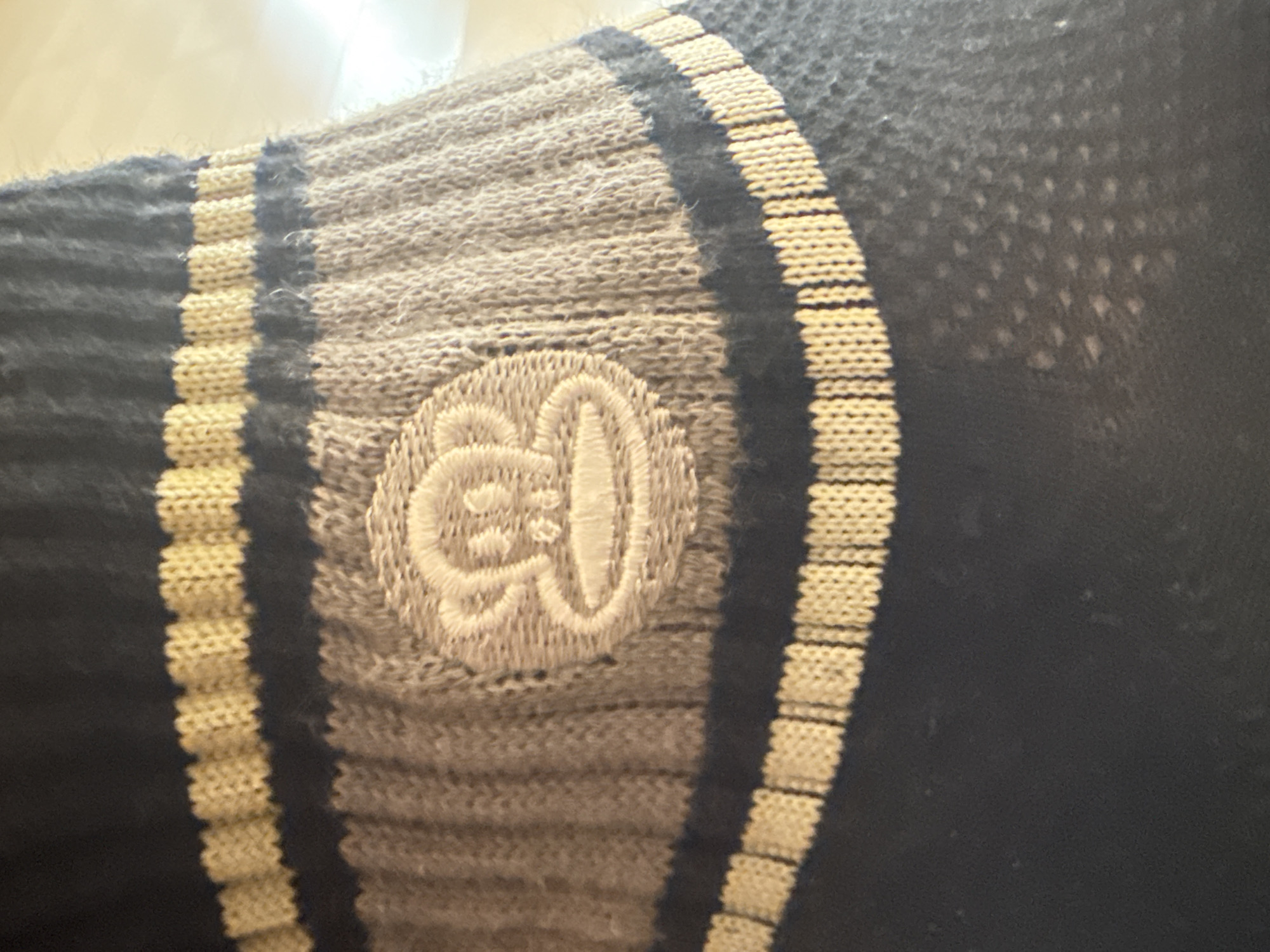 The image displays a close-up of a textile item, likely a sock or a sleeve, worn on a limb. The fabric features a prominent striped pattern composed of varying textures and colors.
The main colors are dark blue/black, light beige/off-white, and a medium grey. The design includes wide dark blue/black sections, interspersed with narrower ribbed stripes of light beige/off-white. A central, wider section is a grey ribbed knit, providing a distinct texture. One of the dark blue/black areas further away from the center displays a subtle, repeating mesh-like pattern of lighter dots, suggesting a breathable fabric commonly found in athletic wear.
A notable detail is an embroidered circular logo or symbol, rendered in off-white thread, situated on the grey ribbed section. This symbol is stylized and appears to be a cartoonish or abstract character, possibly resembling a head with two small eyes and an open, irregular mouth, or a unique letter design. The embroidery features clear outlines and some filled-in areas.
The overall texture of the garment varies from ribbed knit in the lighter stripes and grey section to a smoother knit with a subtle mesh detail in the dark blue/black areas. The lighting is bright and even, highlighting the fabric's details and the embroidery. The background is out of focus and appears bright, suggesting an indoor setting. No specific activity is depicted.