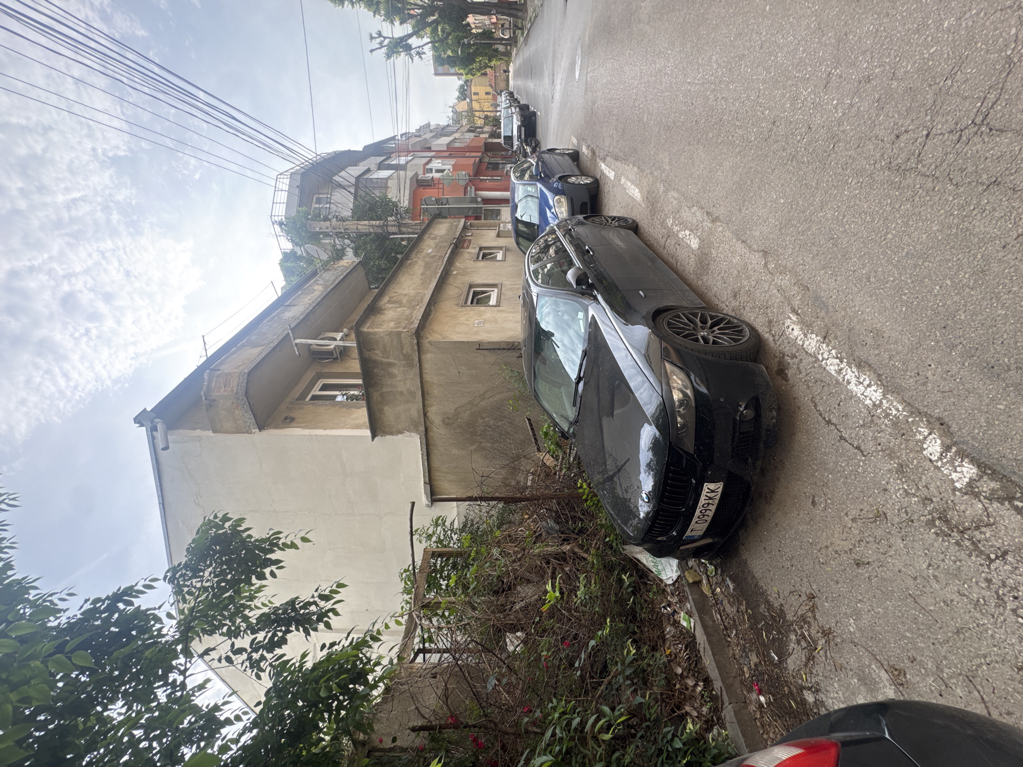 The image captures a narrow residential street in Varna, Bulgaria, under a bright, cloudy sky. The scene is dominated by parked cars and multi-story buildings, suggesting a typical urban neighborhood during the daytime.
In the foreground, a black BMW car is prominently parked on the right side of the asphalt street. Its Bulgarian license plate, "T 0999 KK," is clearly visible, with "T" indicating the Varna district. Adjacent to the car and along the building, there is an unkempt stretch of dense, green bushes and tangled vegetation. Part of another vehicle is visible at the very bottom edge of the frame.
The street itself is asphalt with faded white markings. Further down the road, several other cars are parked in a line, including a blue sedan. On the left side of the street, a row of older residential buildings with varying architectural styles and colors (including grey, white, and reddish-orange facades) line the road. Some buildings feature small balconies, windows, and exterior air conditioning units.
Above the buildings, a network of numerous overhead power lines spans across the bright sky, which shows scattered white clouds. Lush green trees and other foliage are visible throughout the scene, adding natural elements to the urban landscape. No people are visible, and there is no specific activity taking place beyond the stationary cars and the general ambiance of a residential street.