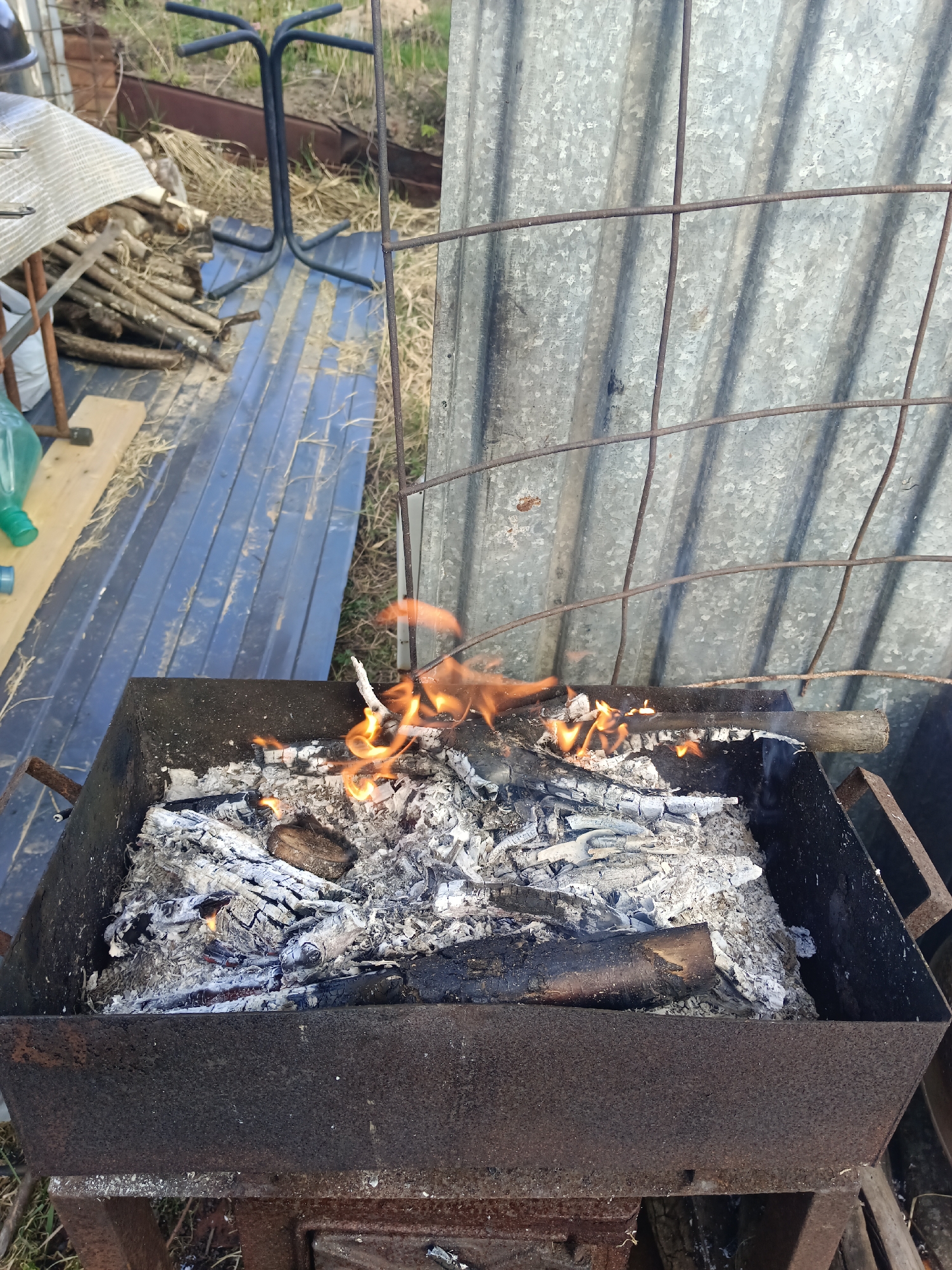 The image shows a metal brazier with burning wood inside. The brazier is mostly filled with ash and embers, but there are still some flames visible. The brazier is sitting outdoors, possibly in a backyard or garden. In the background, there is a metal fence and some other objects, including a table, chair frame and firewood. The location is Вичелово, Russia, based on the prompt.