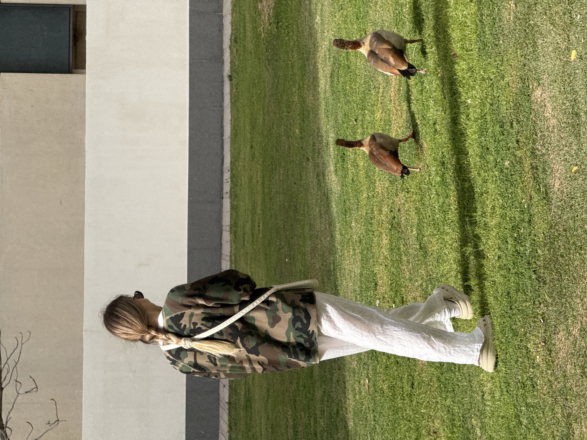 A person with long, light-blonde hair styled in a braid, wearing a camouflage jacket, white pants, and light-colored shoes, is standing on a grassy lawn next to a modern building. Two brown and tan birds are walking on the grass. The photograph appears to have been taken during daylight. The location is likely outdoors, adjacent to an urban building with a lawn. There is no definitive information to confirm if this image was taken in Tel-Aviv or Occupied Palestine.