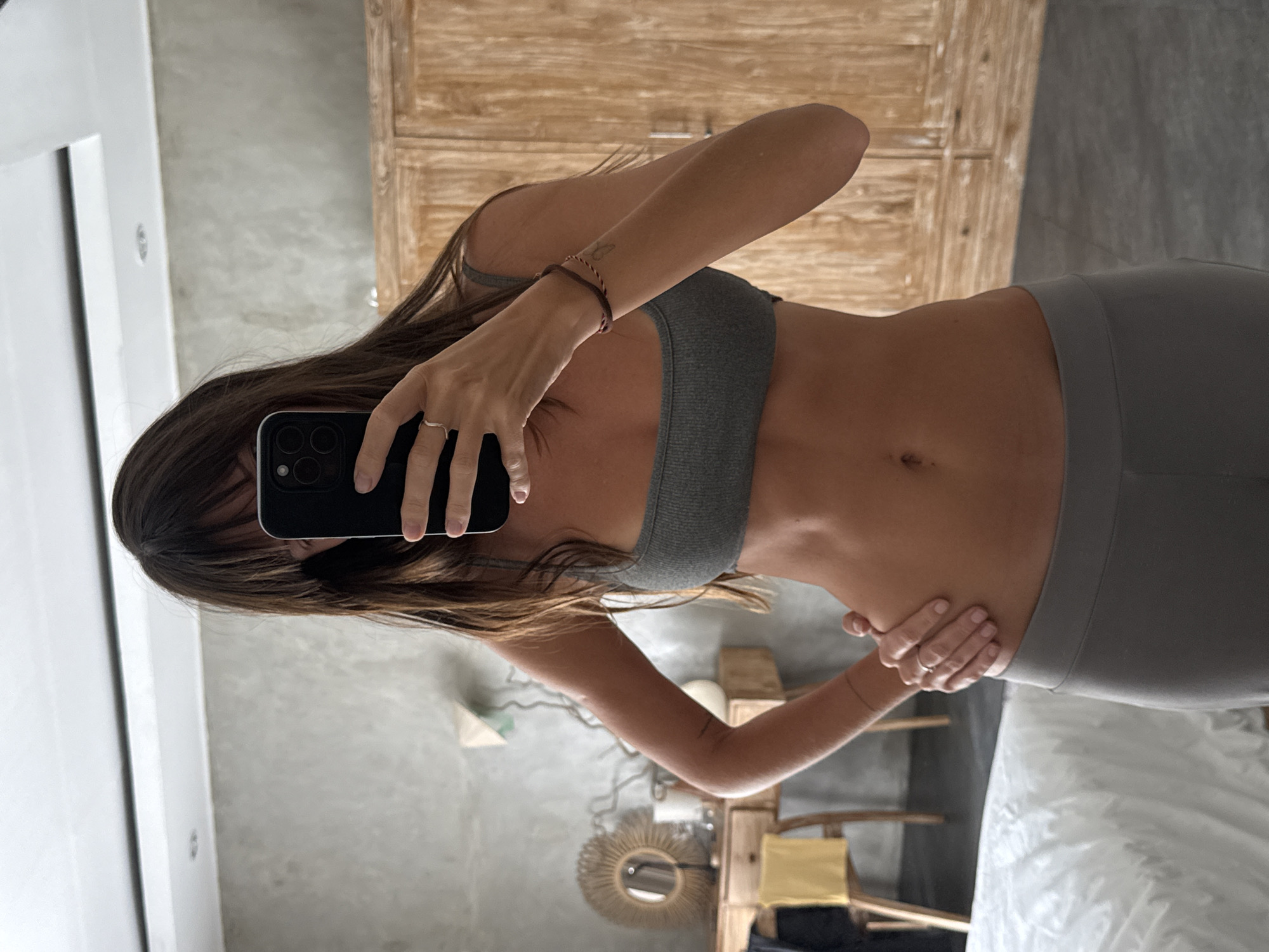 A woman in a gray sports bra and matching leggings takes a mirror selfie. She holds a black iPhone in her left hand, obscuring her face. Her right hand rests on her hip. Her hair is brown. The background includes a white doorframe, gray walls, a wooden cabinet, a decorative wall mirror, and furniture. The location appears to be a room interior, and is speculatively located in North Kuta, Indonesia.