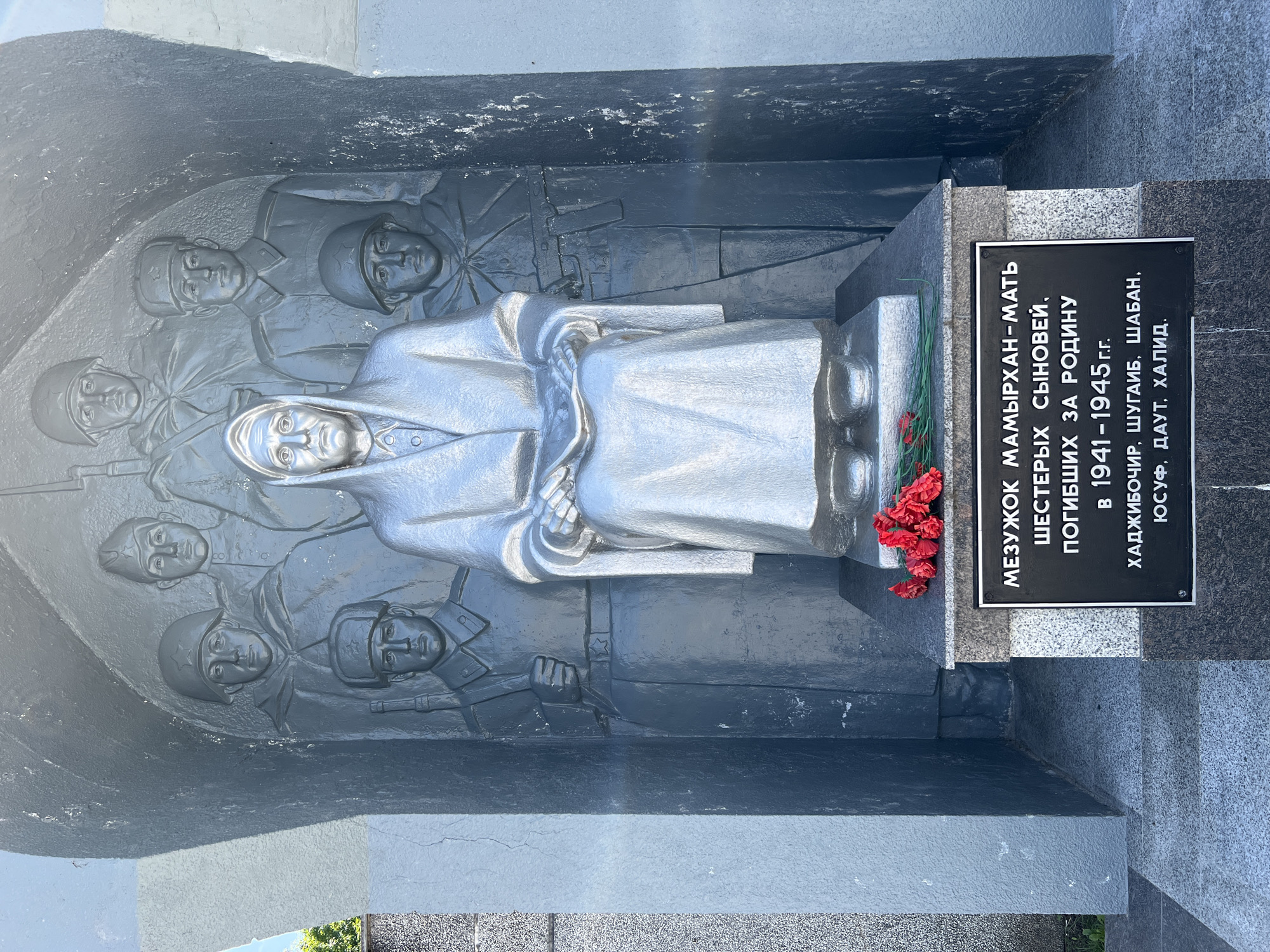 The image shows a monument featuring a central seated female figure made of silver, draped in a robe and head covering, holding an unseen object in her lap. Behind her, a relief sculpture depicts six male figures in military attire. The monument is situated within an alcove, and at its base, a plaque with Russian text is visible, along with a small bouquet of red flowers. The plaque reads "MEZUKHOK MAMYRKHAN - MOTHER OF SIX SONS, WHO DIED FOR THE MOTHERLAND IN 1941-1945 G.G. KHADZHIBICHER, SHUGAYB, SHABAN, YUSUF, DAUT, KHALID." This monument is located in the city of Takhtamukai, Russia. The surrounding environment appears to be an outdoor setting with hints of greenery and sunlight.