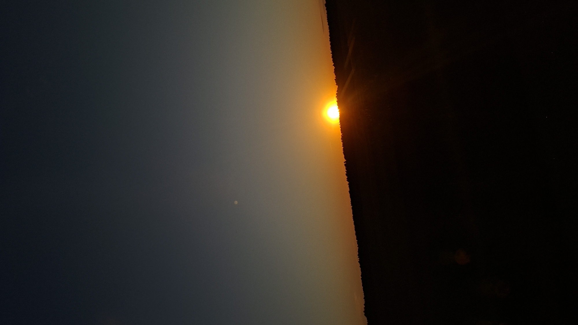 A landscape photograph shows a sunset. The sky on the left is dark blue, fading to yellow-orange in the middle where the sun is setting. The horizon line on the right is dark and indistinct, possibly trees or land. The sun is a bright orange orb. There's a small, pale dot in the sky on the left. The photograph appears to be taken at a tilted angle. The location is Любанскі сельскі Савет, Belarus.