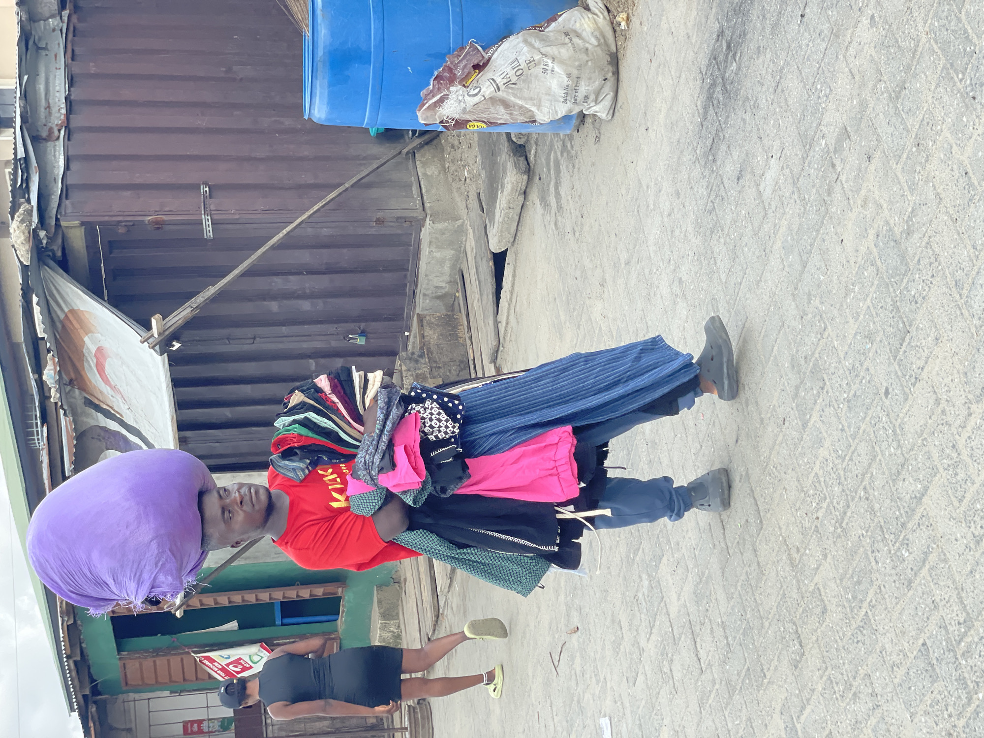 A street vendor, a man, walks along a paved path in an outdoor market or commercial area in Ado, Nigeria. He carries a significant load, balancing a large, round purple bundle on his head and a stack of various colorful clothes in his arms, suggesting he sells garments like skirts, dresses, and tops. He wears a red t-shirt with visible lettering, possibly "KIKI", blue trousers, and dark grey shoes. His expression is neutral, focused on his task.
In the background, another person, a woman, walks away from the viewer. She is wearing a black dress, a black baseball cap, and bright yellow or lime green slip-on shoes.
The setting features utilitarian buildings constructed from corrugated metal panels and shipping containers. A partially visible white and red banner with "airtel" and "HER" is seen on the left. Further back, a blue plastic barrel and a large white sack are present. Text on the sack includes "VOLGA", "50 KG", "Batch No. :- 01", "Date of Prod", "NDIA", and "CE". The ground is paved with light-colored concrete pavers arranged in a herringbone or chevron pattern. The even lighting suggests an overcast day or perhaps early morning/late afternoon, with no harsh shadows. The scene captures the bustling nature of street vending in an urban environment.
