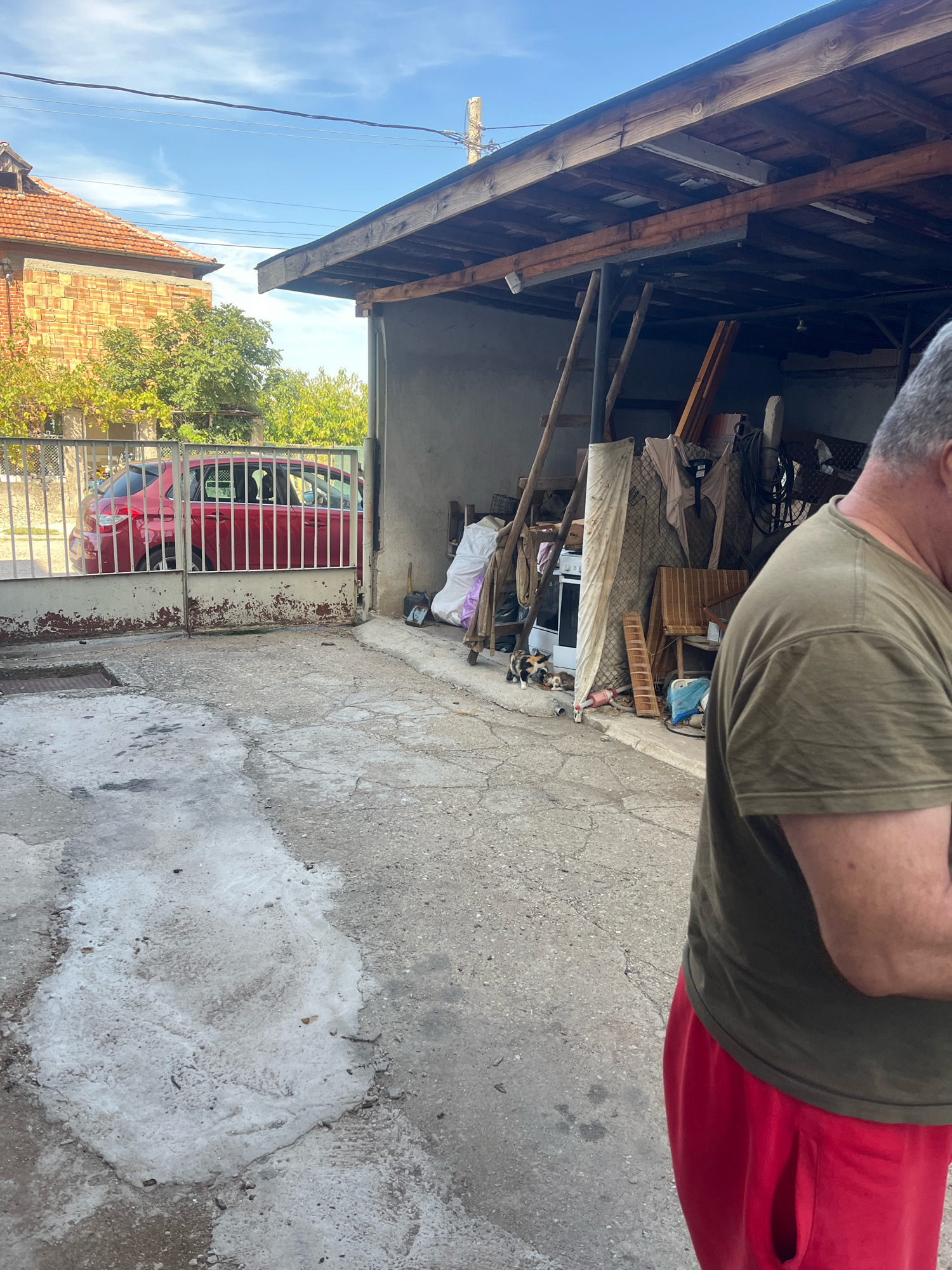 The image depicts an outdoor scene in Cheshnegirovo, Bulgaria. In the foreground, a person wearing an olive green t-shirt and red pants is visible from the shoulders down, with their back turned to the camera. To the left, a red car is parked behind a metal gate. The background features a building with a red tiled roof, a covered structure with a wooden roof, and various stored items including a stove, mattress, and what appears to be a ladder. The ground is paved with concrete, showing signs of wear and discoloration. The sky is blue with scattered clouds.