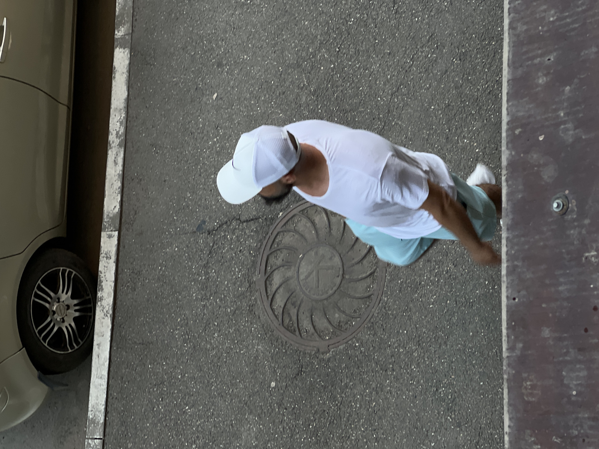 An aerial shot captures a man walking on an asphalt road next to a parked car and a wooden structure. The man, wearing a white t-shirt, a white baseball cap, and light blue shorts, is walking towards the right of the frame. He is looking down, and his right leg is blurred, indicating movement. A manhole cover with a circular, fan-like pattern is visible on the road beneath him. The car, a light-colored sedan, is parked on the left side of the frame. The wooden structure, appearing to be a wall or fence, is on the right edge of the image. The ground is asphalt speckled with small white stones. The lighting suggests it might be daytime, possibly overcast. The scene appears to be an urban or suburban environment. There is no visible text in the image.