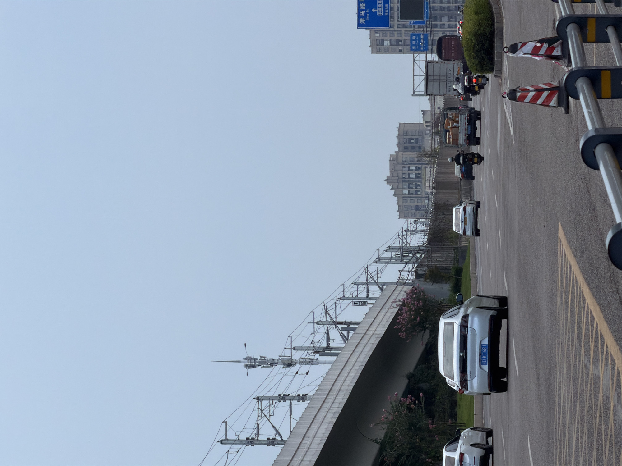 In the city of Jiangjin, China, on a cloudy day, an overpass carrying train tracks dominates the left side of the frame, with utility poles and wires stretching across the sky. A light blue sedan with a license plate ending in "681" is driving on the road in the foreground. In the background, a white sedan and multiple motorcycles are also visible on the road. A truck is visible to the right, carrying boxes. Buildings with a Brutalist architectural style are visible in the distance. Traffic cones and a metal railing line the side of the road.