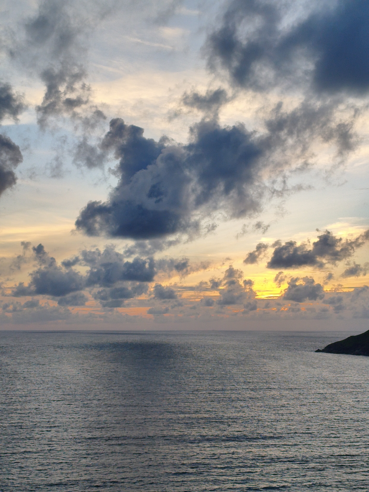 This is a photograph of a seascape at sunset. The sky is filled with dramatic clouds, ranging from dark gray to lighter shades of blue and pink. The sun is partially visible, casting a bright yellow glow through a gap in the clouds. The ocean is calm with a rippled surface, reflecting the colors of the sky. In the distance, on the right side of the frame, a dark green headland juts out into the water. The scene appears to be captured from an elevated vantage point, looking out over the sea towards the horizon. This is likely a location in Rawai, Thailand, known for its picturesque coastal views, especially during sunset. There are no people or boats visible in the image.