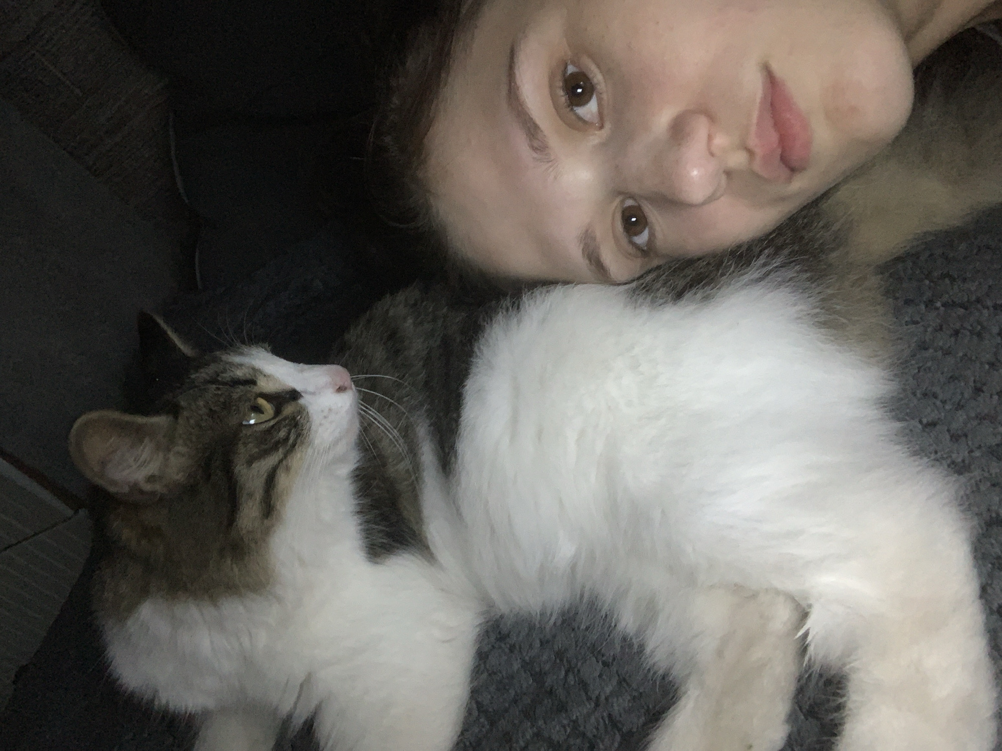 In Belogorsk, Russia, a close-up, high-angle shot captures a young woman and a cat. The woman, on the right, has brown eyes and light skin, with her face angled towards the camera. Next to her, the cat, with white and tabby markings, is lying down, facing away from the woman and looking to the left. The background is dark and indistinct, suggesting an indoor setting. The composition emphasizes the shared space and proximity between the woman and her pet.