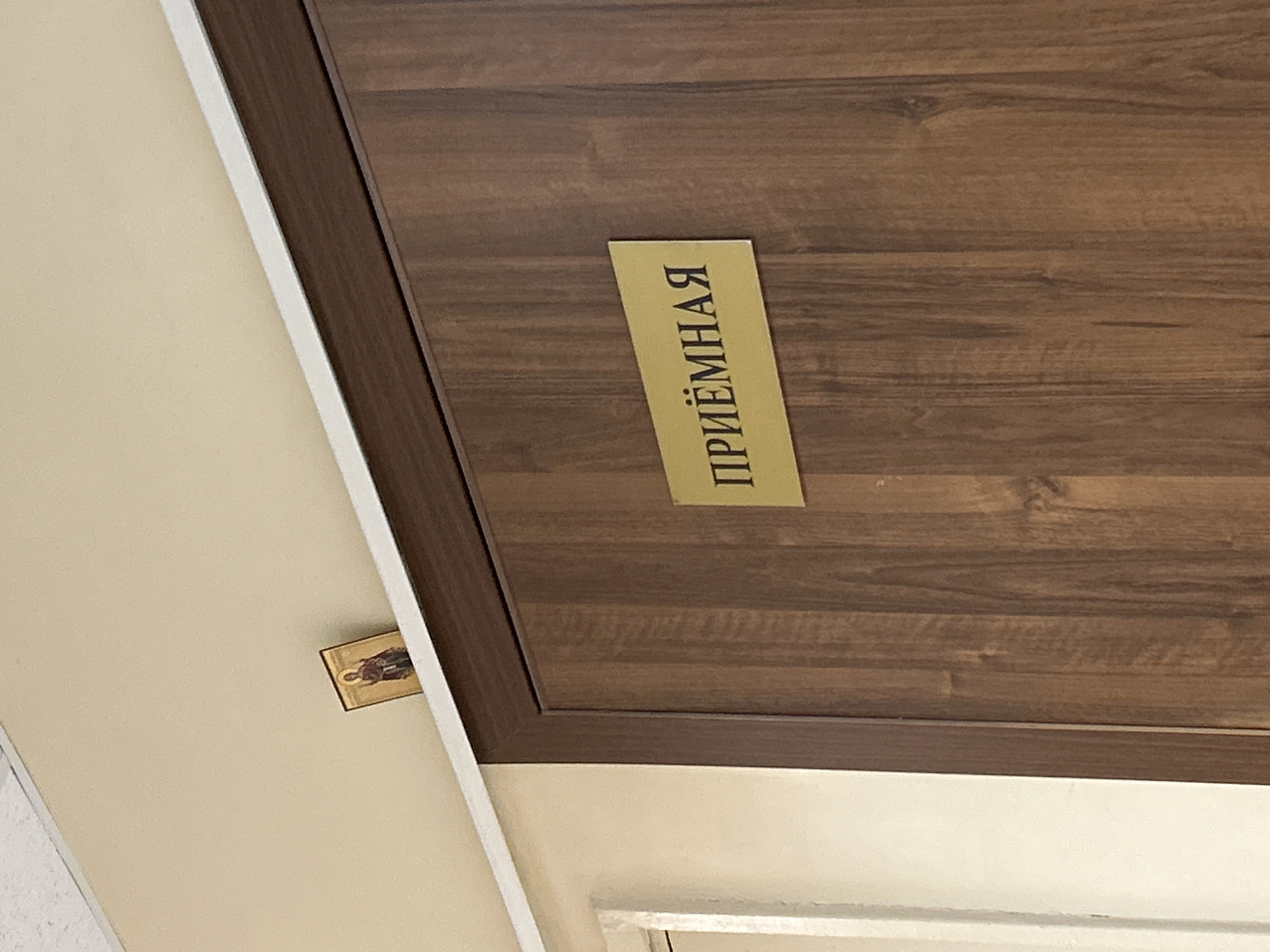 The image shows a wooden door with a yellow sign that reads "ПРИЁМНАЯ" (Reception). Above the door, on a ledge where the wall meets the ceiling, rests a small, ornate religious icon. The icon depicts a figure in traditional religious attire. The overall scene appears to be inside a building, possibly an office or a public institution. The location is identified as Lyubertsy, Russia.