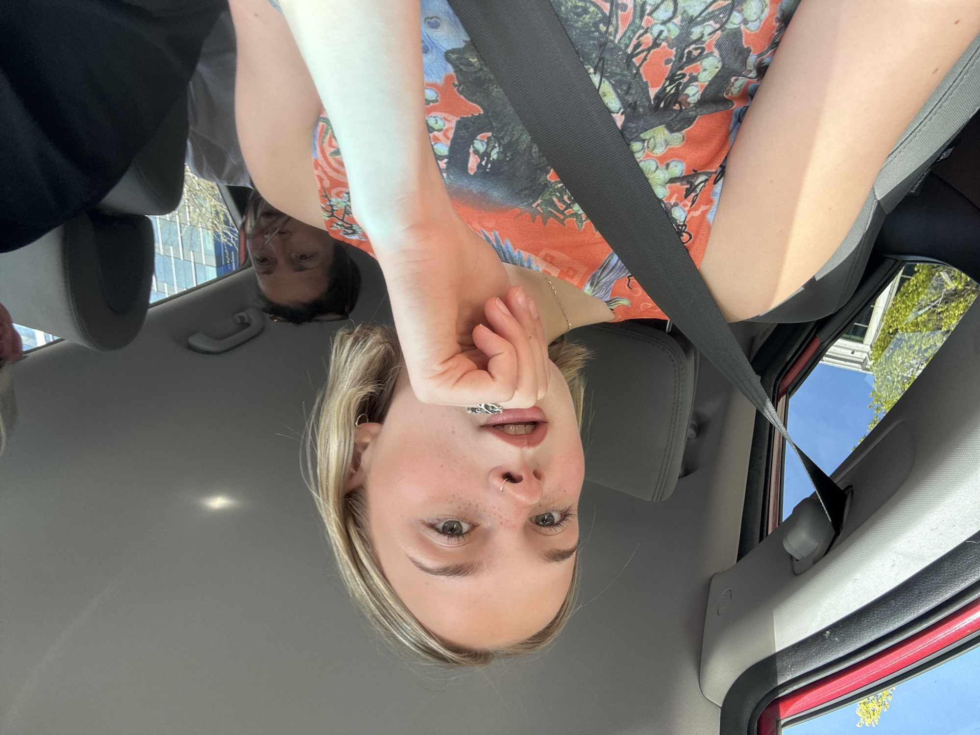 A photograph, oriented upside down, shows the interior of a car. The main subject is a fair-skinned individual in the passenger seat, looking directly at the camera with a hand near their face; they have blonde hair, green eyes, and a nose ring. They are wearing a floral patterned top and a seatbelt. Another person, presumably the driver, is partially visible in the background. The car's interior is visible, including the headrest, ceiling, and part of the dashboard. Outside the car window, buildings and greenery are visible, likely indicating an urban setting. Given the request for the location, and based on visual clues, the photograph likely taken in Kelowna, Canada.