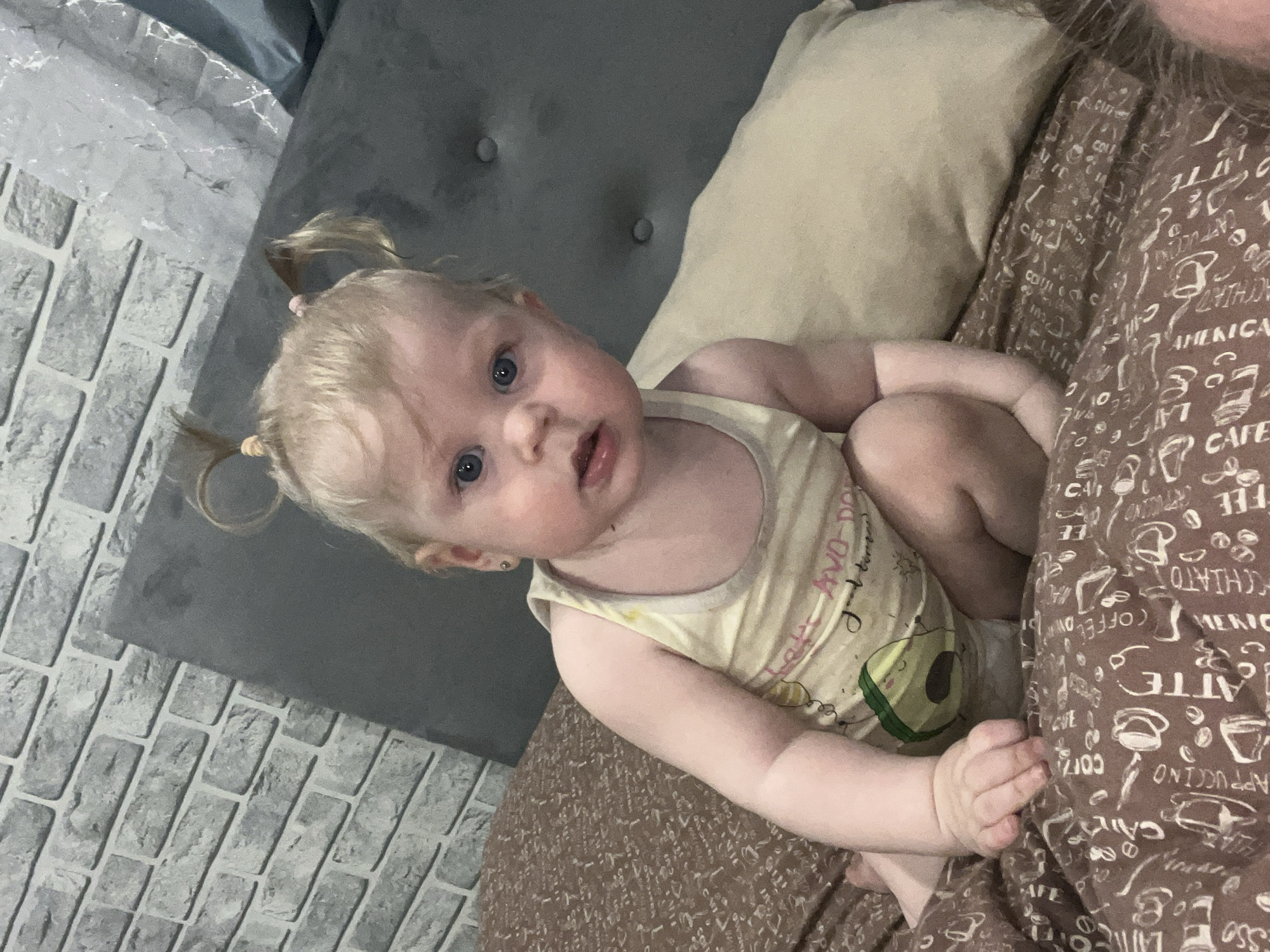 A baby with blonde hair styled in pigtails is lying on a bed. The baby is wearing a yellow tank top with an avocado graphic. The bed has a beige pillow and a brown patterned blanket with coffee-related words. The background includes a gray headboard with buttons and a gray brick-patterned wall. The scene appears to be indoors. There is no indication or mention of the city Vesyoloye, Russia, in the image.