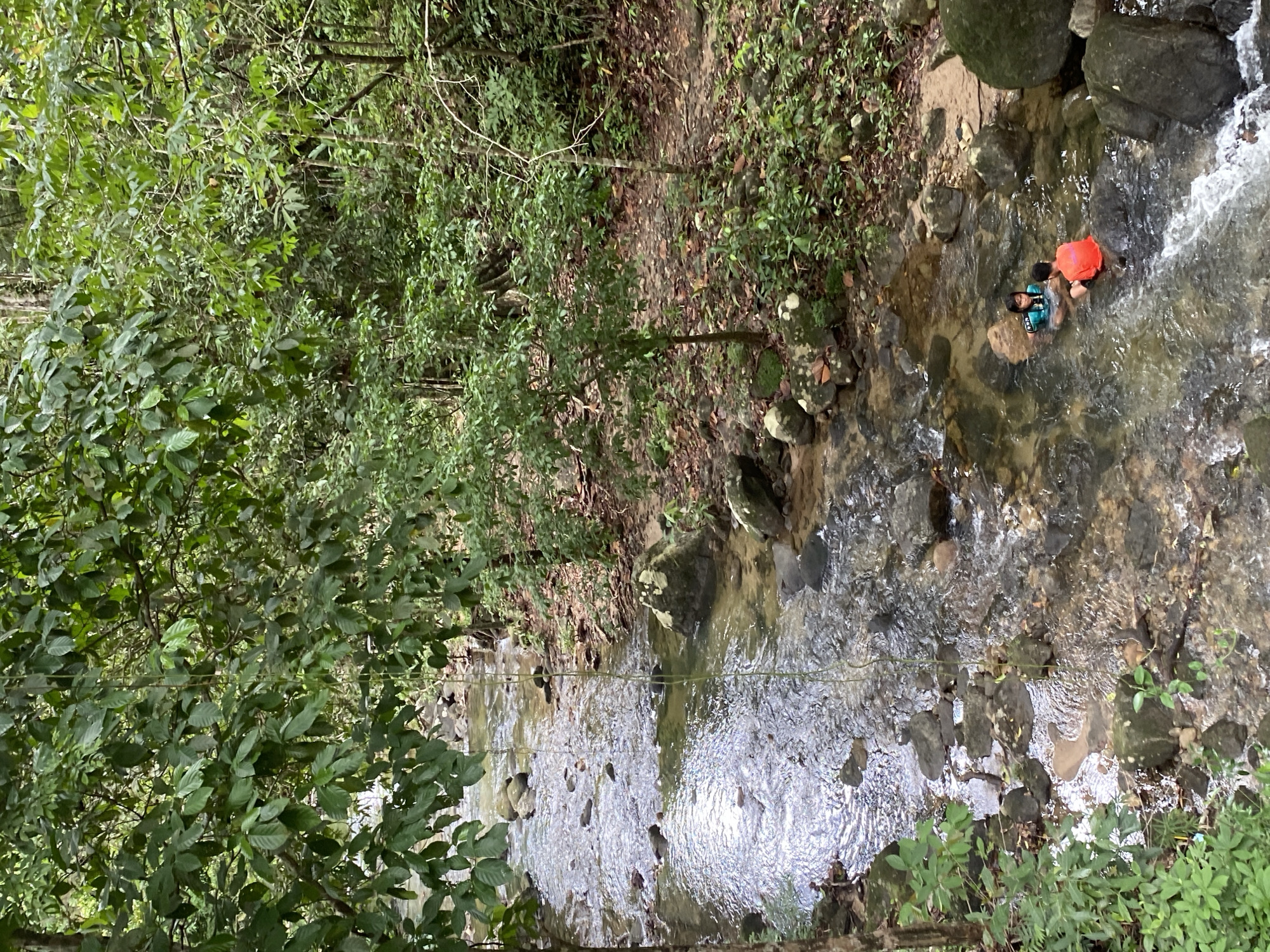 Two people are in a shallow, rocky river in Tambon Thai Mueang, Thailand. One person wears an orange shirt and the other wears a blue and green shirt. The river flows through a dense forest. The scene is viewed from an elevated position, looking down.