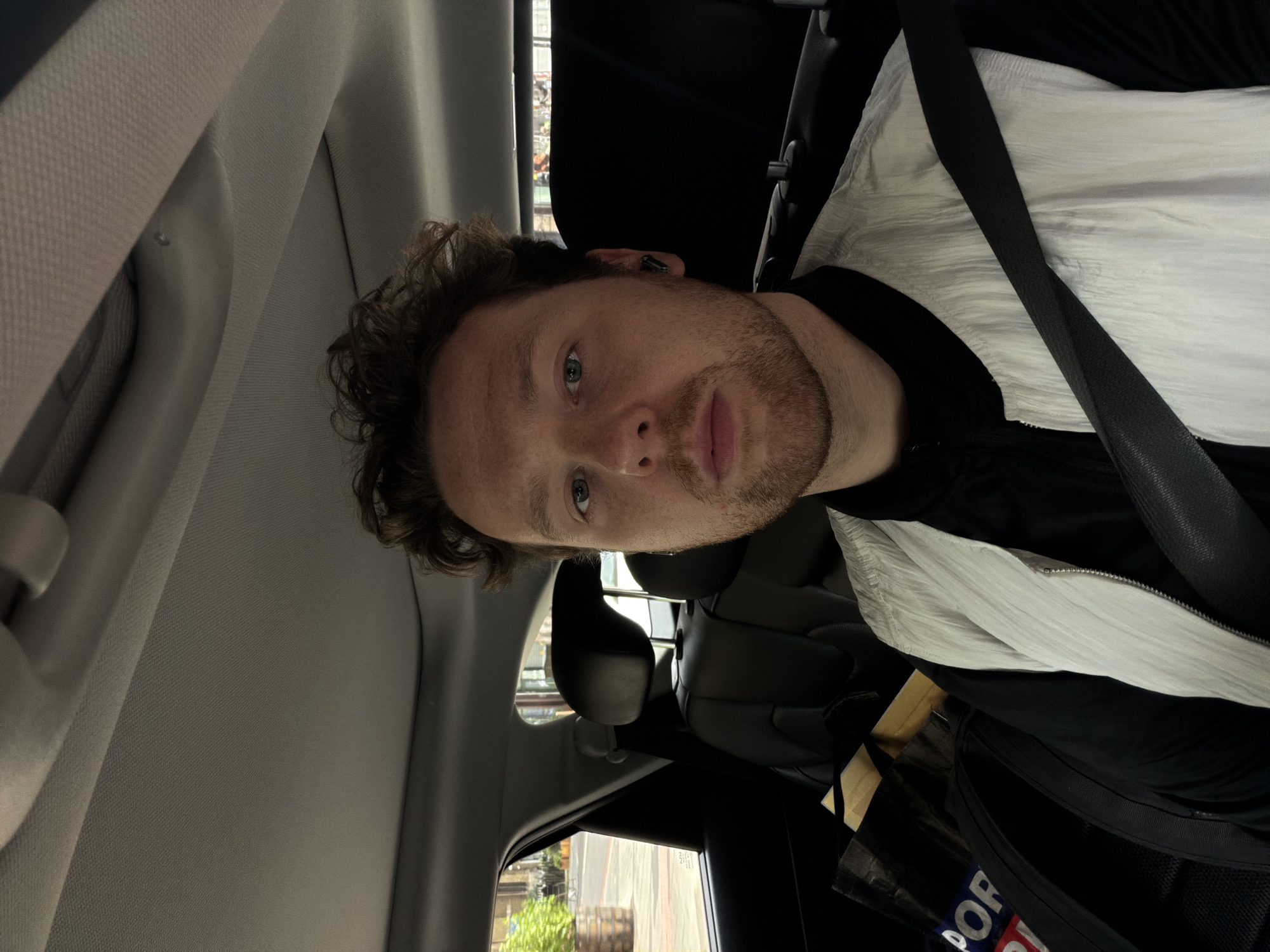 A man with curly brown hair and a light beard is in the passenger seat of a car, wearing a black shirt and a white jacket with a black seatbelt across his chest. He is looking directly at the camera. The car's interior is visible, including the seat, headrest, and side panel. The background shows a street scene outside the car window, with buildings and greenery, possibly in the city of London, United Kingdom.