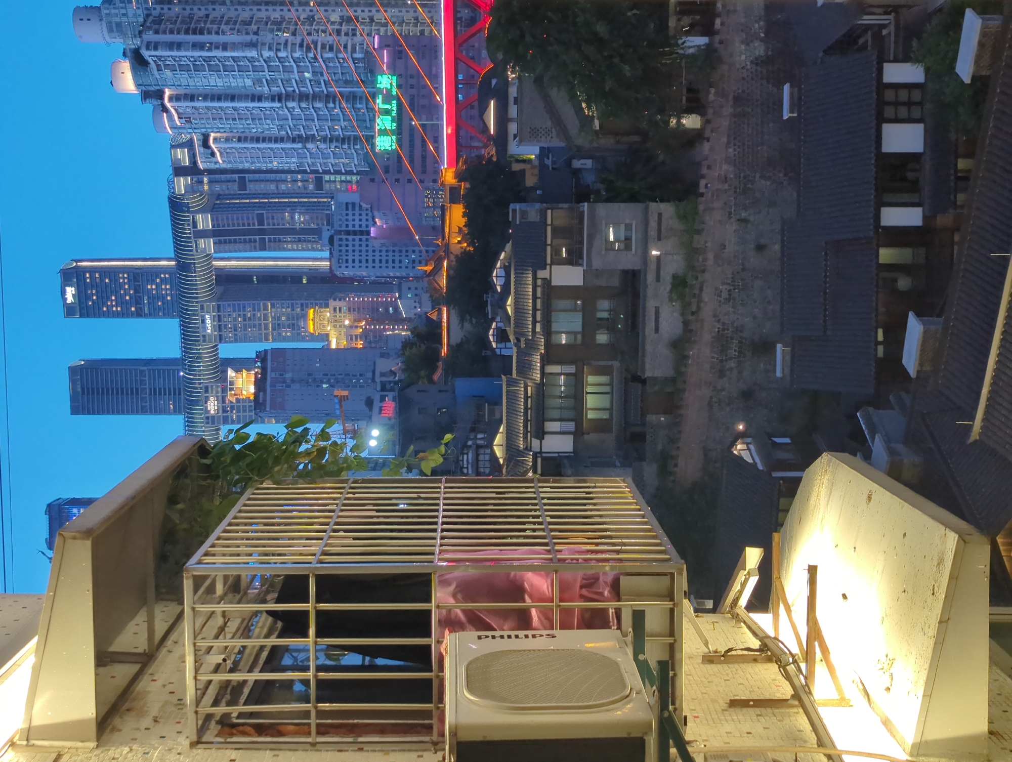 The photograph depicts a twilight scene in Yuzhong District, China, capturing a layered urban landscape. In the foreground, a white air conditioning unit is mounted on a textured white wall, partially obscured by a metal grate. The grate encloses a pink fabric material. Above the AC unit, a beige awning with attached lighting casts a warm glow downwards.
The midground reveals a dense collection of buildings with traditional tiled roofs, some featuring illuminated windows. A stone wall with crenellations separates these lower structures from the buildings behind.
The background is dominated by modern skyscrapers, some connected by an elevated, illuminated walkway. These tall buildings are lit from within and by external lighting, creating a vibrant cityscape against the darkening blue sky. A prominent red bridge structure is visible in the far right, with orange lights tracing its outline. Green Chinese characters on a building in the distance read "港口广场" which translates to "Harbor Plaza". The overall atmosphere suggests a bustling urban environment transitioning from day to night.