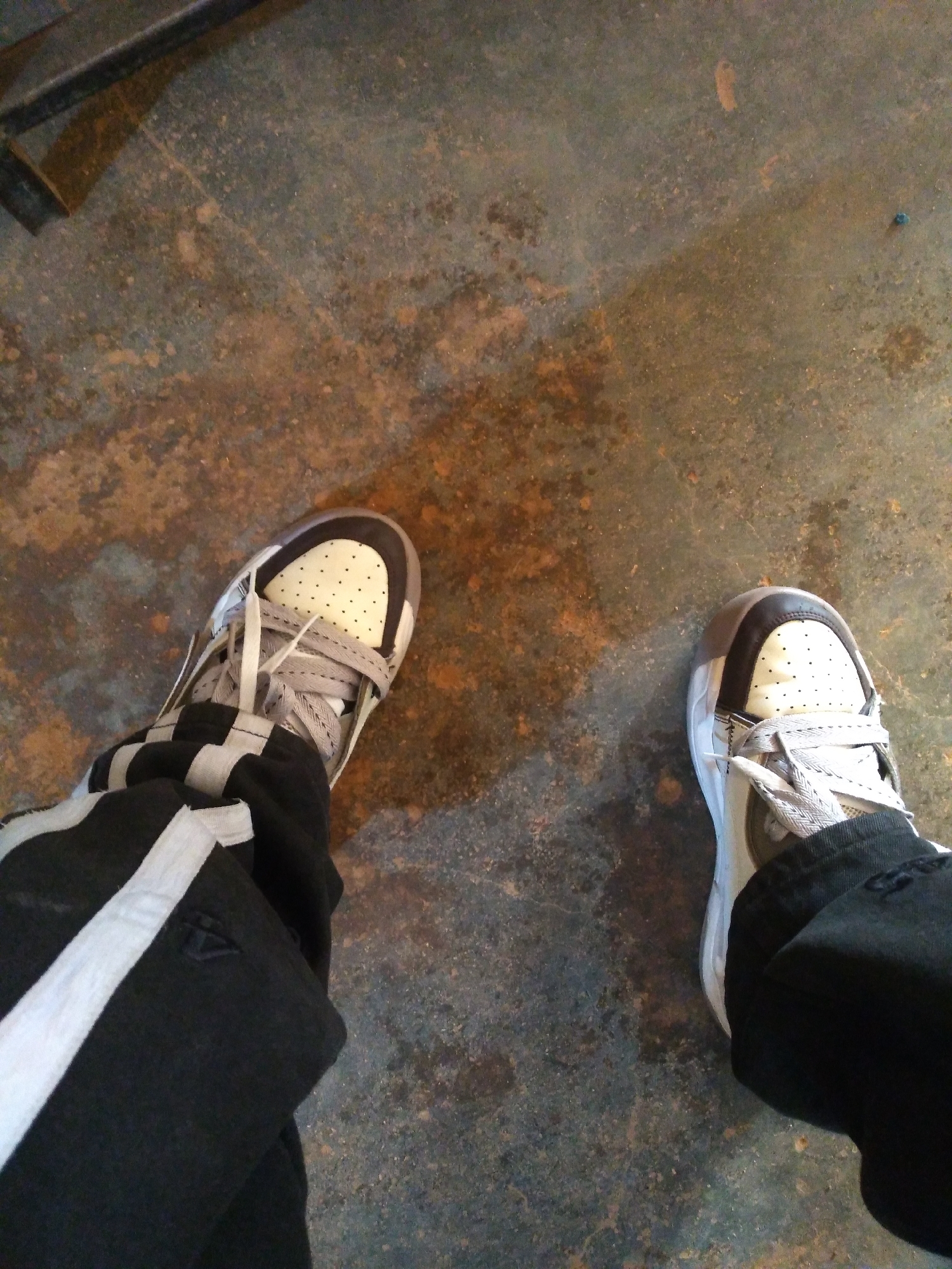 A pair of feet wearing black pants with white stripes and white and gray sneakers are standing on a dark, textured concrete floor. The floor has patches of rust and discoloration. A dark, metal object is partially visible in the upper left corner of the image. The scene appears to be indoors, possibly in a workshop or industrial setting. No individuals are fully visible, only the lower legs and feet. The location is not identifiable as Jos, Nigeria, as there are no specific visual cues to indicate this.