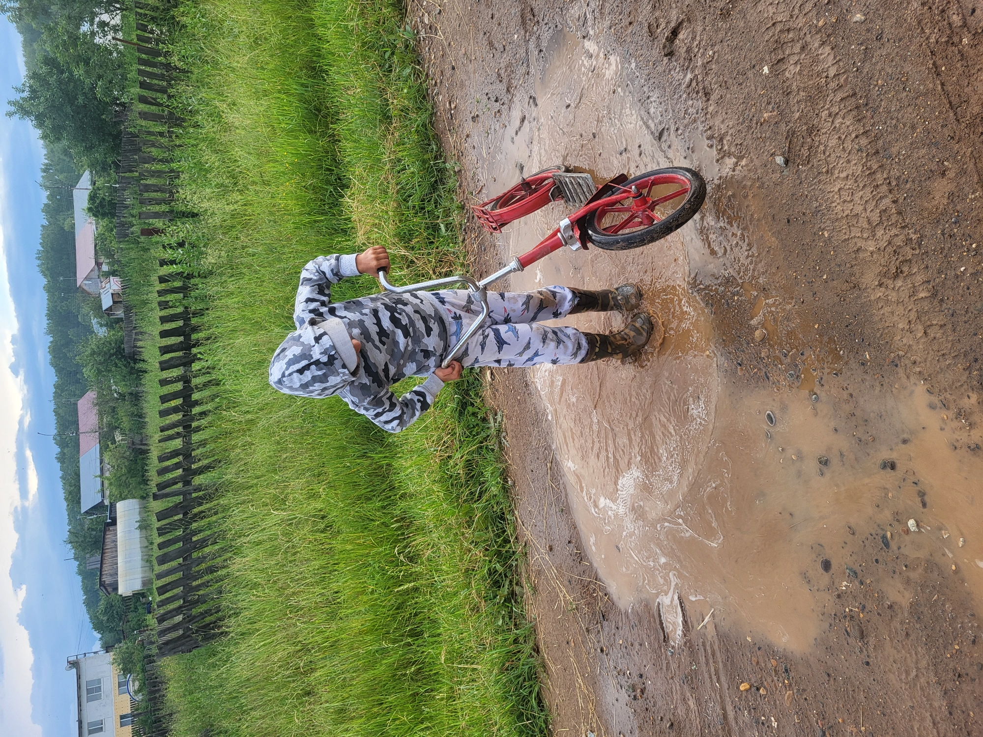The image shows a person in a camouflage outfit standing in a large puddle of water on a dirt road. The person is holding onto the handlebars of a red bicycle that is lying on its side in the puddle. The person is wearing tall black boots. The scene is outdoors, with a grassy area to the left and a wooden fence running along the edge of the grass. In the background, there are houses and trees, suggesting a rural or village setting. The sky is overcast, indicating it may be a cloudy or rainy day. The dirt road is muddy, with tire tracks visible. The image appears to be taken in daylight. The overall impression is of a child playing in the mud and puddles. No visible text is present in the image.