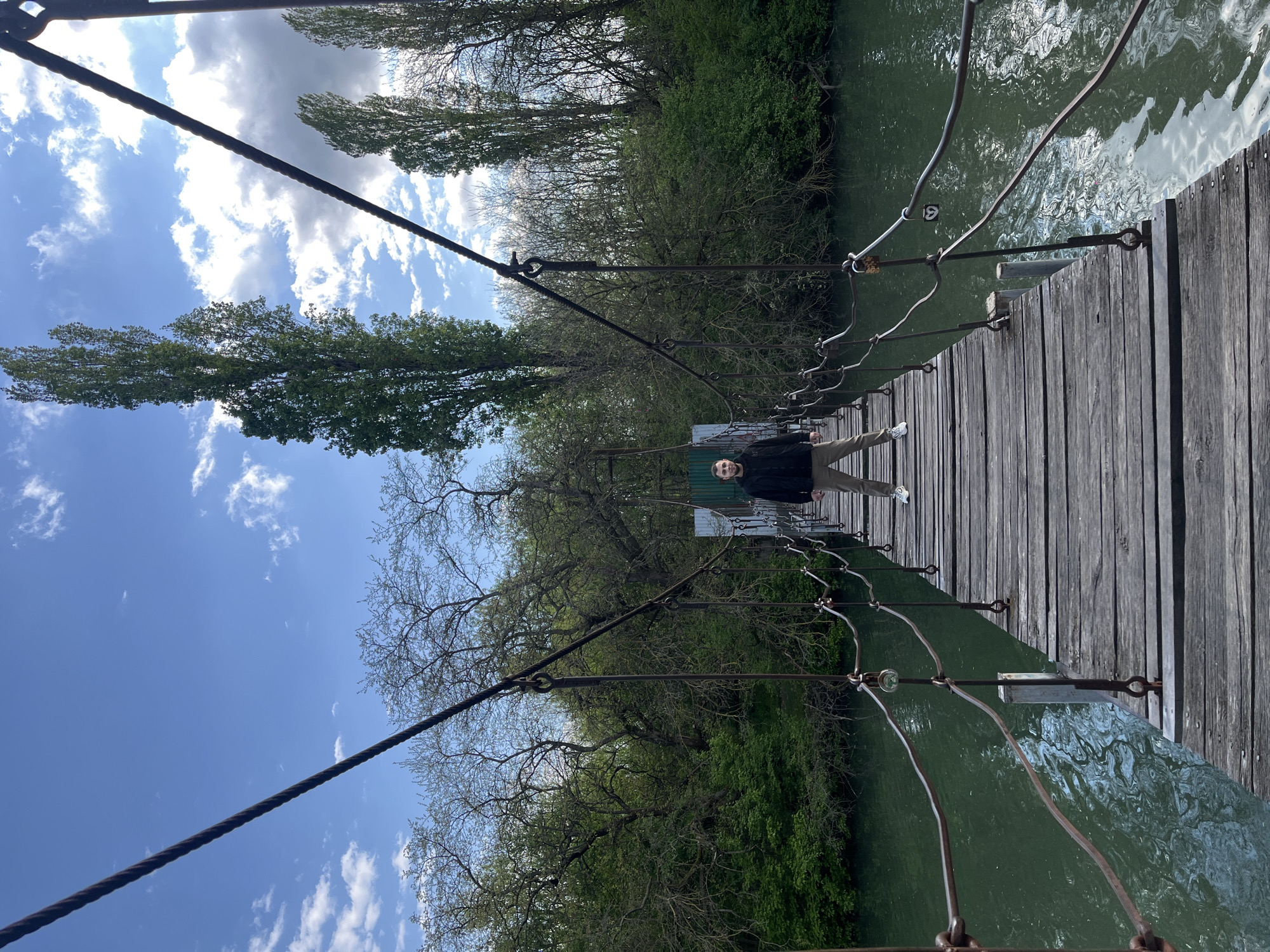 The image depicts a person standing on a wooden suspension bridge over a river. The bridge is constructed with wooden planks, metal cables, and supports. The person is wearing a dark jacket and light-colored pants. The river flows beneath the bridge, and trees line both banks. The sky is visible above the trees, with some clouds. The image may have been taken in the city of Krasnodar, Russia.